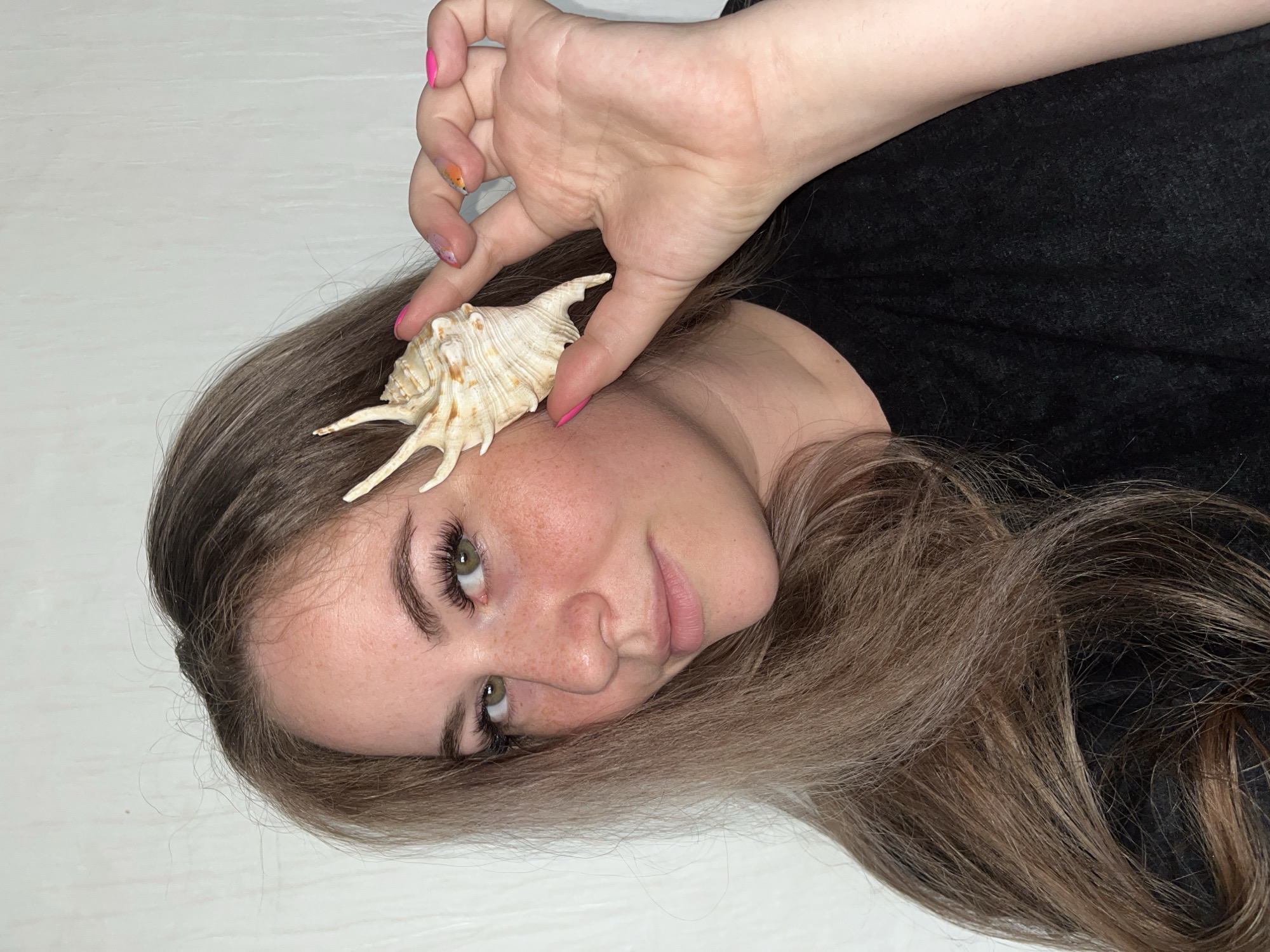 A young woman with long brown hair is holding a seashell near her face. She is wearing a black shirt and has colorful nail polish. The background appears to be a white surface.
While it is not possible to confirm from the image, there is no information or indicators that would identify this location as Сосново, Russia.