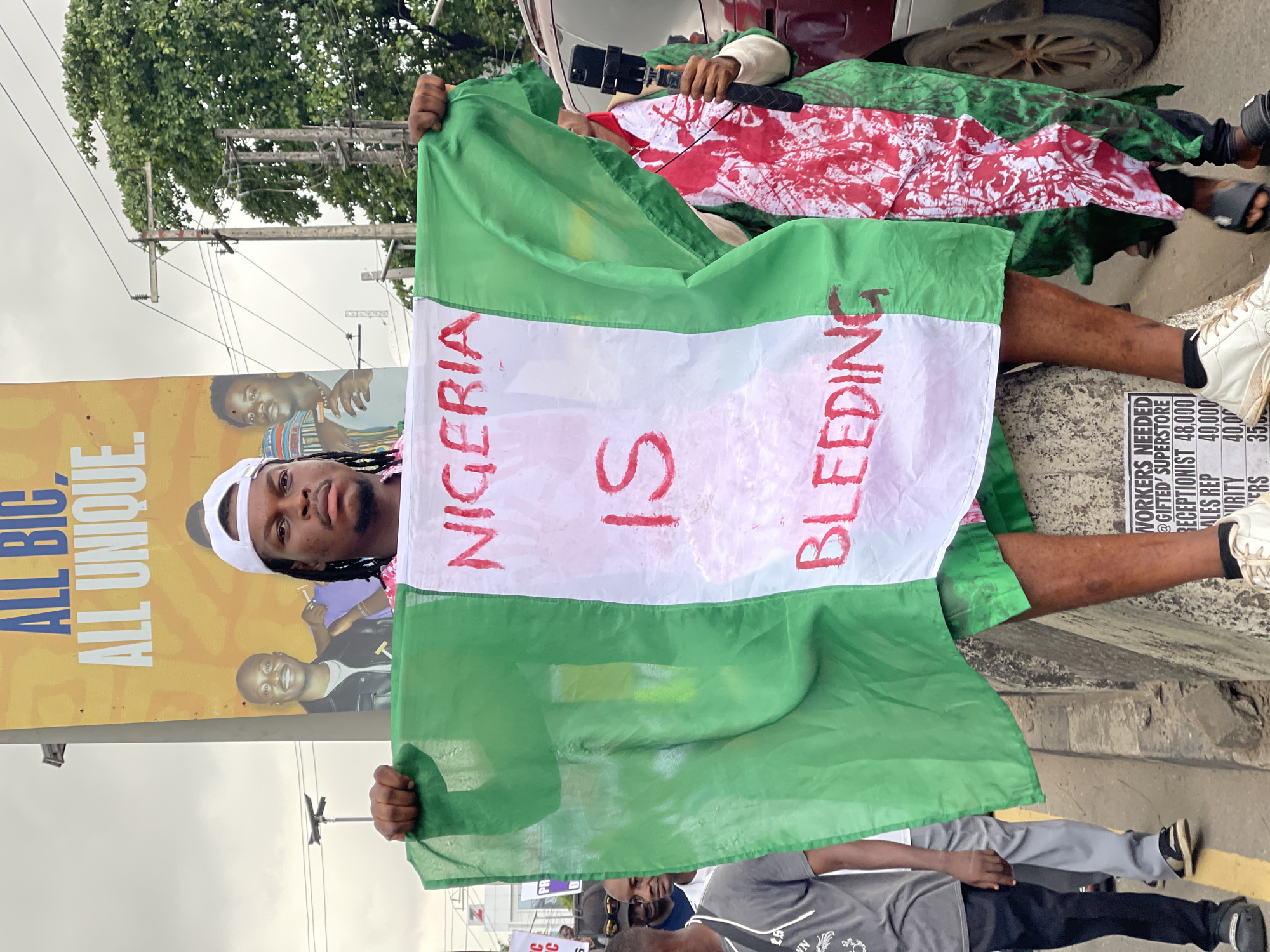 In Ikeja, Nigeria, a young Black man wearing a white baseball cap, a pink patterned shirt, and green shorts is holding up the Nigerian flag. The flag has the words "NIGERIA IS BLEEDING" written in red paint on its white stripe. He is seated on a concrete barrier, and his legs are bare. Behind him, a billboard displays text including "ALL BIG, ALL UNIQUE" and images of other Black men. In the background, other people are visible, as well as trees, power lines, and vehicles, suggesting an outdoor urban setting. The scene appears to be a protest or demonstration.