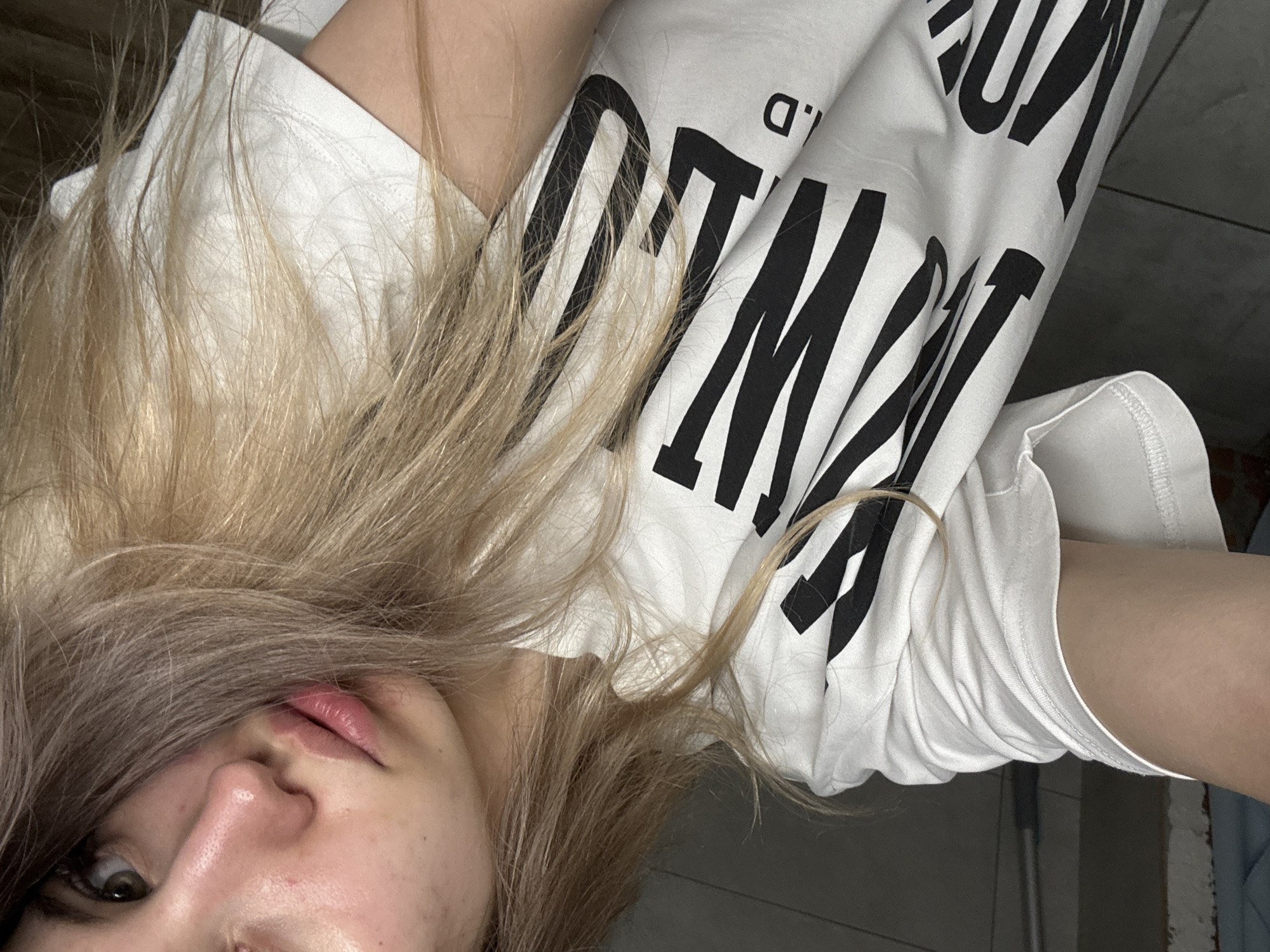 A person lies on a floor in an indoor setting, wearing a white t-shirt with large black printed text that includes ".D". The person's face is partially visible, showing a light skin tone, brown eyes, and blond hair that transitions to brown at the roots. The floor appears to be tiled and the overall lighting is somewhat dim. No explicit location or specific actions can be determined, however, the location is mentioned in the query to be Chaikovsky, Russia.