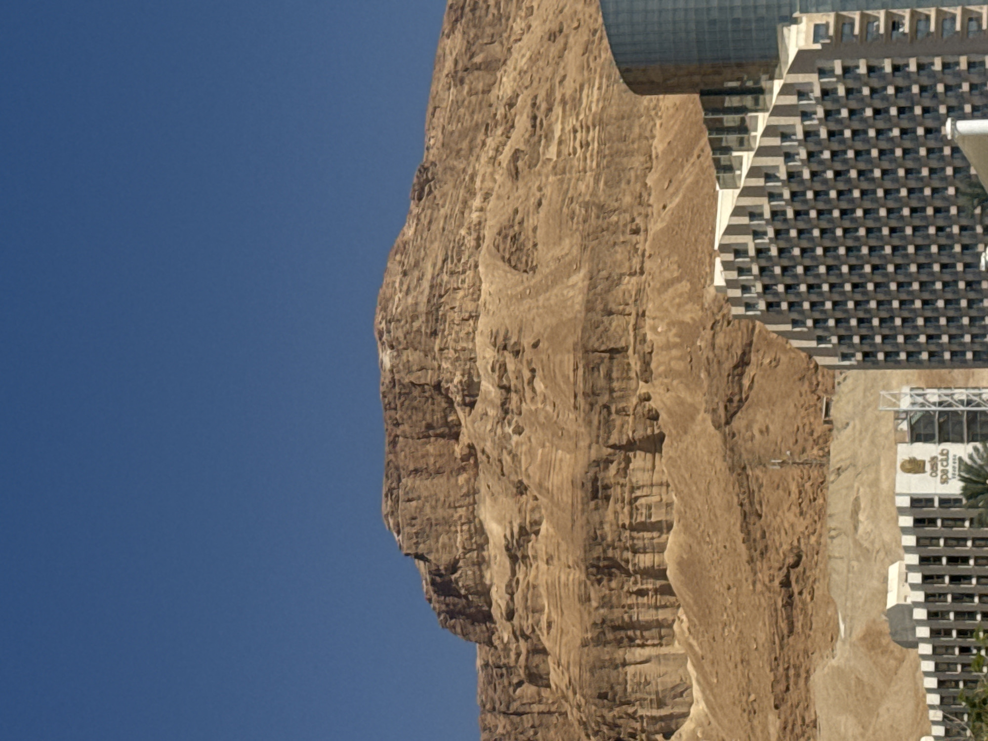The image shows a view of a rocky cliff face alongside a modern building. The cliff is tall and textured, appearing naturally weathered with various shades of brown. A clear blue sky occupies the left side of the frame, contrasting with the cliff's earth tones. To the right, a large, multi-story building with a geometric pattern of windows and balconies is visible. The building's facade appears to be a mix of concrete and glass, giving it a contemporary look. Palm trees are visible near the base of the building. A closer inspection reveals a spa logo on the side of the building "Oasis Spa Club". The location is Tamar Regional Council, Israel.