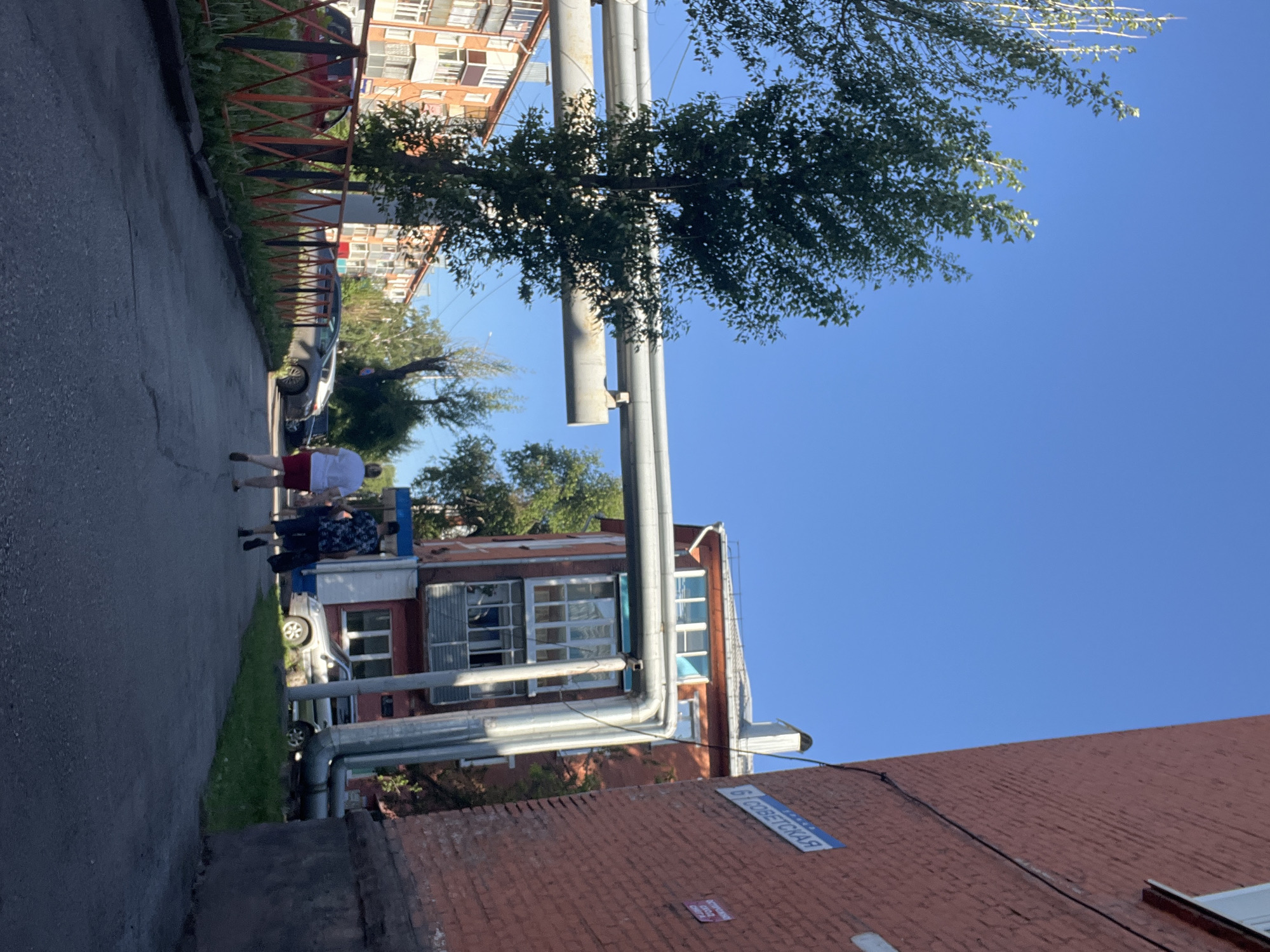 The image depicts a street scene in the city of Kiselyovsk, Russia, under a clear blue sky. A red brick building, part of a larger apartment complex, is on the right side of the frame, with a street sign labeled "6 СОВЕТСКАЯ" (6 SOVIETSKAYA) attached to its wall. To the left, another apartment building with multi-colored balconies is visible. A network of large, grey pipes runs horizontally across the middle of the image, supported by brackets on the red brick building. In the foreground, two individuals are walking away from the camera down an asphalt path. One person is wearing a white t-shirt and red shorts, and the other is wearing a patterned shirt and dark shorts, carrying a black bag. Several cars are parked on the left side of the path, behind a low, orange and black metal fence. Trees with green foliage are present on both sides of the scene.