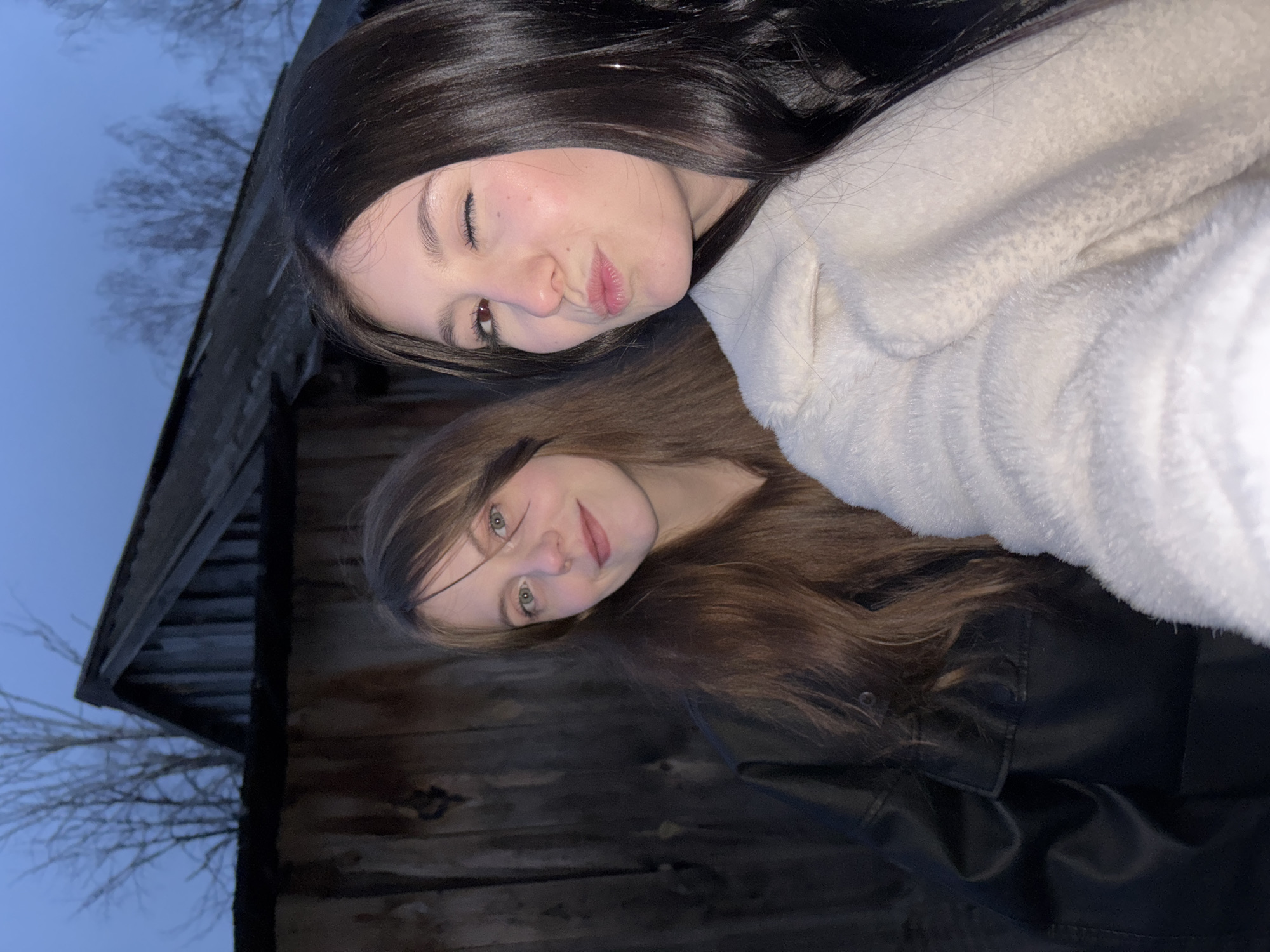 Two young women pose for a selfie outdoors near a wooden structure, possibly a shed or outbuilding. The woman at the top of the frame has dark hair and wears a light-colored, fuzzy jacket. She has her eyes closed. The woman below has brown hair and wears a black jacket. A leafless tree is visible against the sky. The setting appears to be Фоки, Russia.