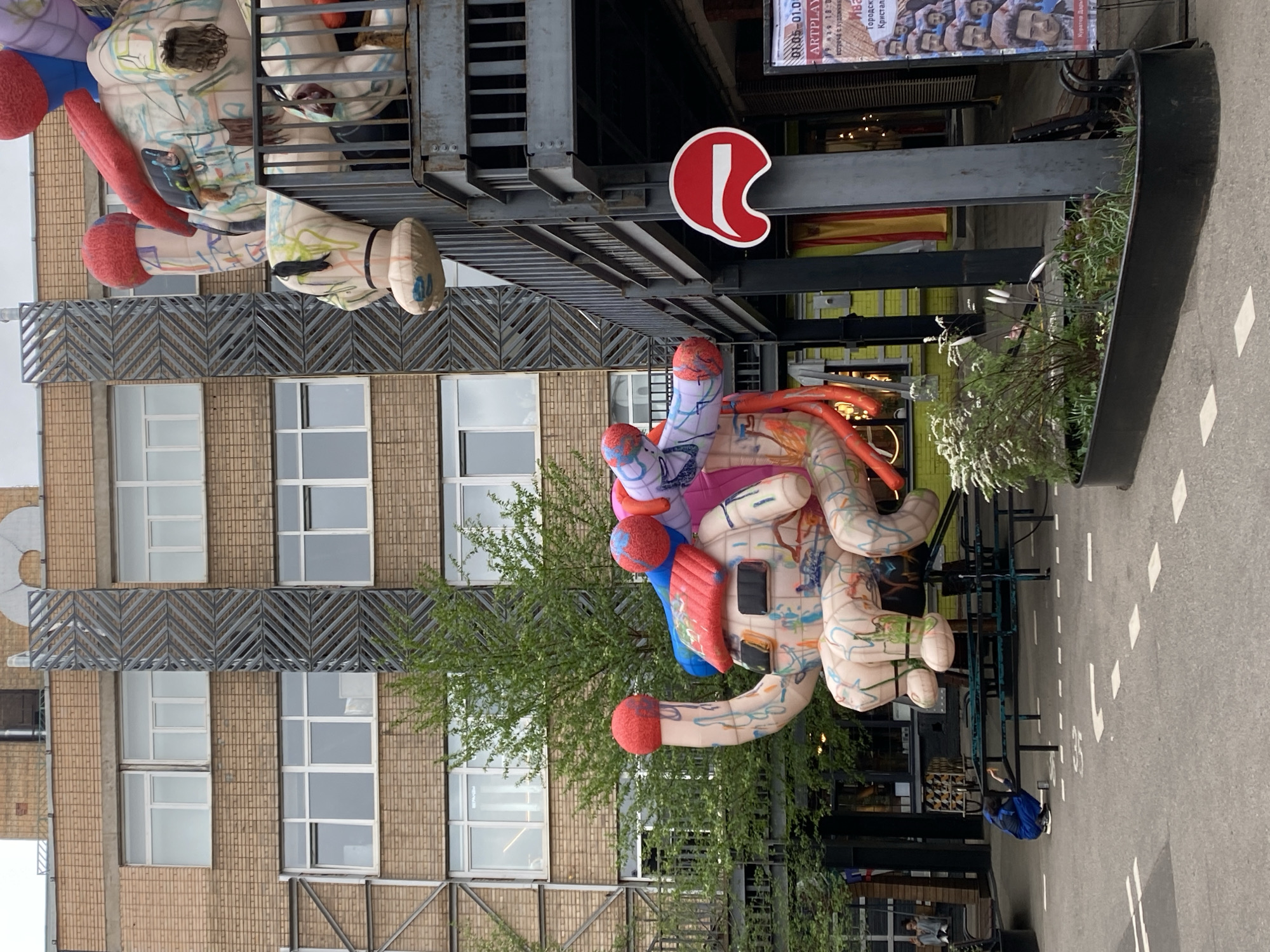 The image shows a street scene in Moscow, Russia. An inflatable sculpture, brightly colored and abstract in form, hangs from a building's facade. The building is multi-story with a brick exterior and gridded windows. A metal walkway is visible, extending from the building's exterior. Below the sculpture, there's a street or open space. Seating areas are apparent, and a person wearing a blue jacket is kneeling. A red and white logo is attached to the building. A planter filled with greenery is adjacent to the seating. A poster is visible on the building with pictures of various people.