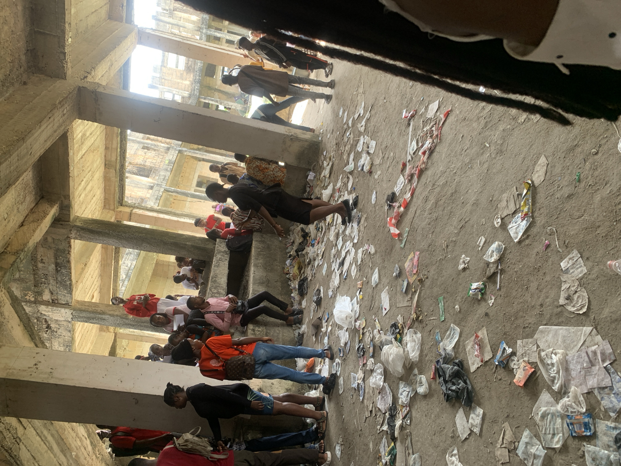 The image depicts approximately 25-30 individuals, predominantly young adults, congregated within the ground level or an open-air floor of a large, unfinished or derelict multi-story concrete structure in Uyo, Nigeria. The environment is characterized by exposed concrete pillars and beams supporting a raw concrete ceiling. The dirt and concrete floor is heavily littered with various items, including numerous plastic bags, food wrappers, paper fragments, and a visible strip of red and white striped barrier tape.
Individuals are observed standing, walking, and sitting on a low concrete ledge or elevated platform. Their attire is casual, comprising t-shirts, jeans, shorts, and footwear such as sandals or sneakers; several individuals are carrying backpacks or shoulder bags. Bright natural light enters the space through large openings between structural elements, revealing further unfinished or dilapidated concrete buildings in the background. No distinct organized activity or interaction is evident among the individuals; the scene suggests a casual gathering or waiting area. The lower right foreground is partially obscured by the dark dreadlocks and shoulder of an unseen individual.