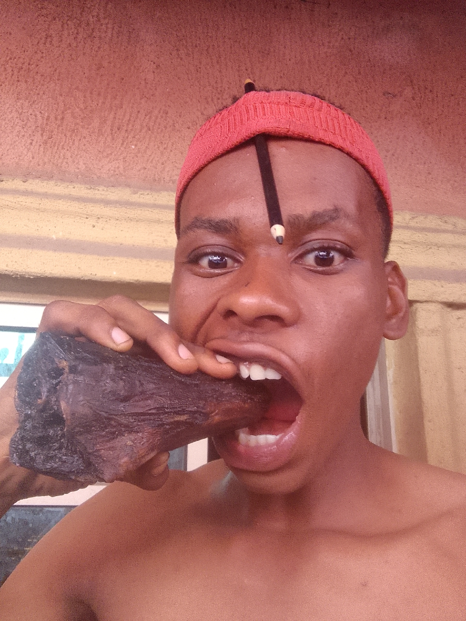 A shirtless dark-skinned person with a red headband and a pencil stuck vertically to their forehead is holding a large, dark, irregular-shaped piece of meat to their mouth. The person's mouth is open, and their teeth are visible. The background is blurry, but includes a light-colored wall on the right and a window on the left. Based on the prompt context, this image is said to be taken in Nkpor, Nigeria.