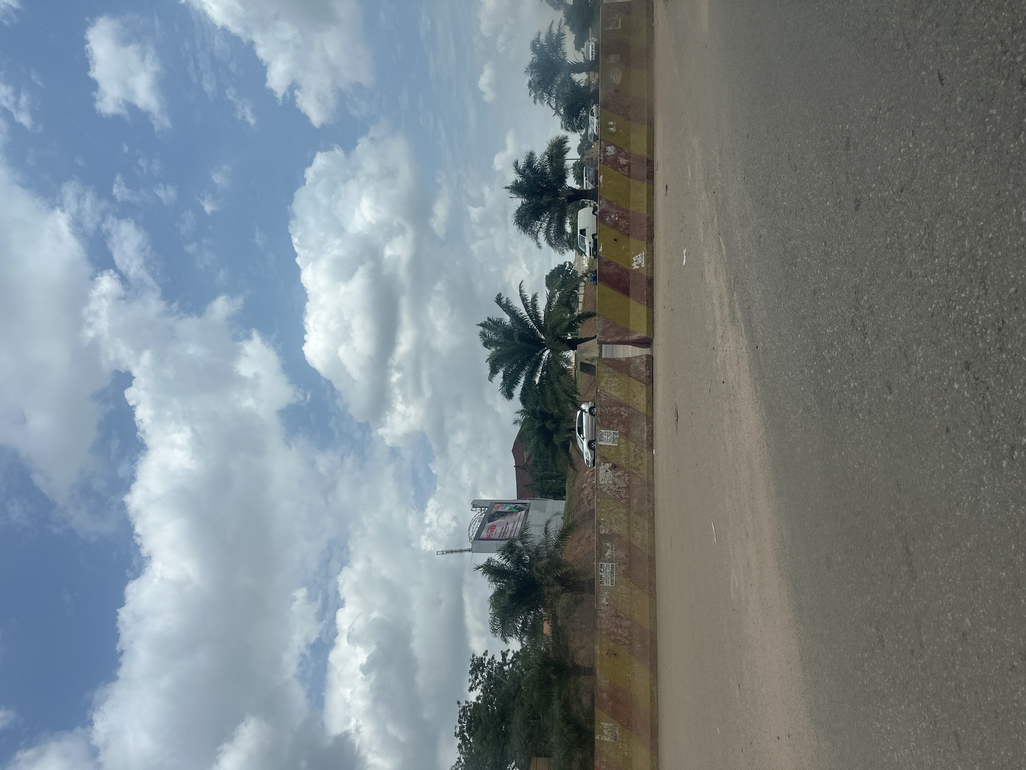 The image captures a street scene in Abuja, Nigeria, under a partly cloudy sky. A concrete barrier, painted with alternating yellow and red diagonal stripes, runs horizontally across the foreground, separating the road from the area behind it. Several vehicles, including a silver car and a white van, are visible on the road behind the barrier. Palm trees are interspersed with other greenery, suggesting a tropical or subtropical environment. In the background, a large billboard stands prominently, displaying the word "POWER" along with some graphics. Buildings and more trees are also visible in the distance. The road surface in the foreground appears to be asphalt, with some faint markings. The overall impression is of a typical urban roadside with signs of ongoing development or infrastructure work.