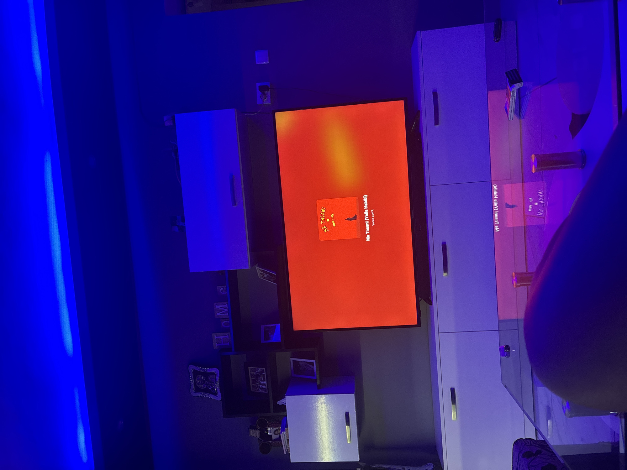The image depicts a dimly lit indoor residential scene, likely a living room, located in Radievo, Bulgaria. The primary light source is overhead blue LED strip lighting, casting a strong blue hue throughout the upper portion of the room, on the walls, and reflecting off surfaces.
In the center of the frame, a large flat-screen television is displaying a vibrant red and orange screen with an album cover and text. The album cover, visible on the left side of the screen, features a silhouette of a person and the title "MA THSANI" with "VANCO H AYA" below. Below the album cover, larger text reads "Ma Tnsani (Yalla Habibi)" and "Vanco H AYA".
Below the television is a white media console or TV stand with multiple cabinets, each featuring a horizontal silver handle. Above and to the left of the television, a wall-mounted shelving unit is visible. This unit contains various decorative items, including framed pictures, a framed icon or artwork in a silver ornate frame, and light-colored letters spelling "HOME". To the far left, a white wall-mounted cabinet is visible, also with a horizontal silver handle. Another small decorative item resembling a Mickey Mouse figurine is on a shelf further left.
In the foreground, a glass coffee table reflects the blue lighting from above and the red/orange light from the television screen. On the coffee table, a clear cylindrical object with an orange top and a rectangular item (possibly a pack of cigarettes or similar) are visible. A black remote control or small electronic device is also on the table. A human leg, clad in dark fabric, occupies the lower portion of the foreground, resting on what appears to be a sofa or chair.
The walls are painted a dark color, appearing blue-purple due to the ambient lighting. Electrical outlets are visible on the wall to the right of the television. Wires are partially visible behind the upper right cabinet.