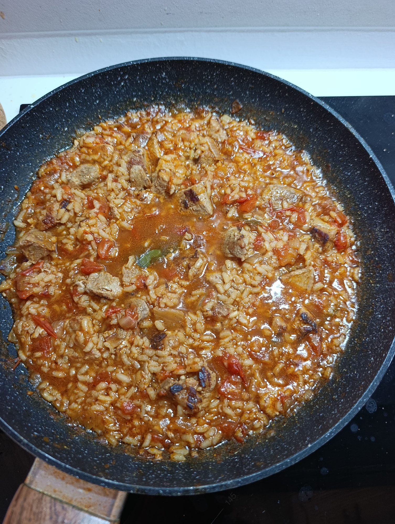 This is a top-down, medium shot of a paella dish being cooked in a dark grey, speckled frying pan. The paella appears to be made with rice, diced tomatoes, and pieces of meat. The dish is saucy and glistening, indicating it is actively cooking or has just finished. The pan is resting on a black stovetop. The background is blurred, showing a white countertop and wall. No people are visible in the image. There is no information in the image that indicates the location in the city of Setúbal, Portugal.