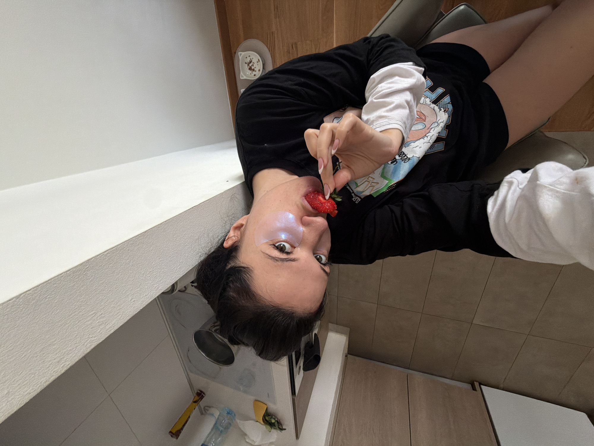 A young woman with dark hair and fair skin is indoors, holding a strawberry to her mouth. She is wearing a black long-sleeved shirt over a white long-sleeved shirt and black shorts, and she is sitting in a gray chair. A clear gel patch is under her left eye. A small white bowl with dark speckles sits on a surface near her head. The background shows a kitchen or similar area, with light-colored cabinets, a countertop, tile flooring, and a trash can. The scene appears to be photographed from a high angle, slightly rotated clockwise. There is no indication of the location being in the city of Belgrade, Serbia.