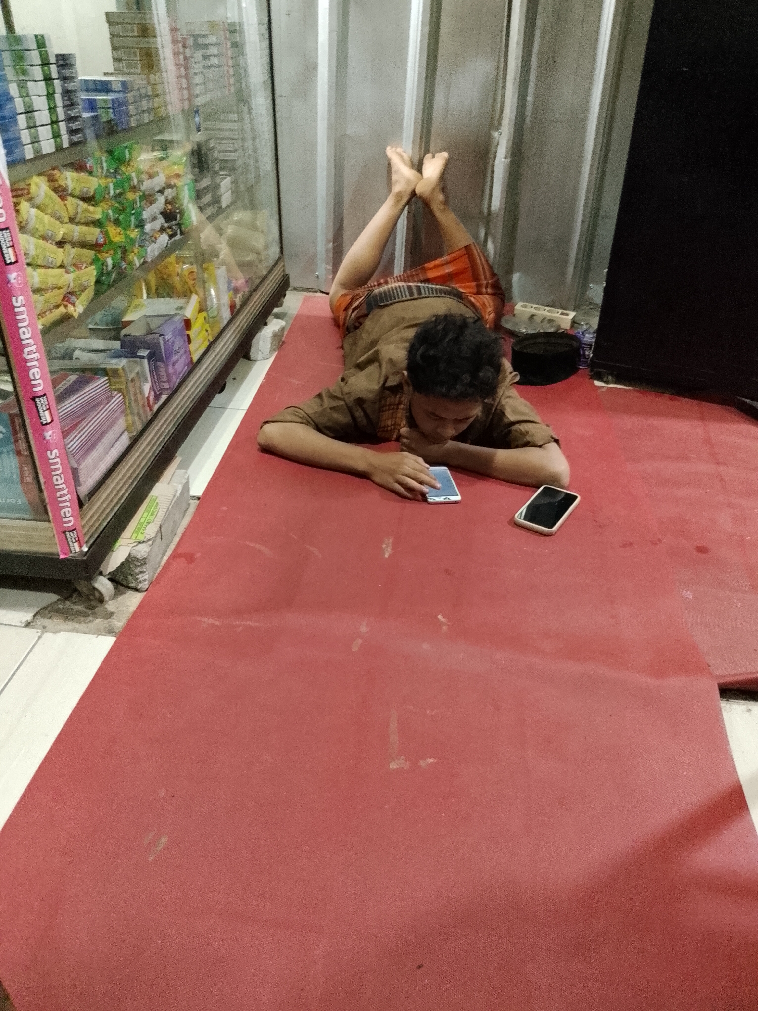 A young male is lying on a red carpet on the floor in a shop in Sukamantri, Indonesia. He is wearing a brown shirt and an orange and brown sarong. His legs are bent and resting against a corrugated metal wall. He is looking down at two cell phones on the floor in front of him. One phone is white and the other is black. Behind him, to his left, is a glass display case filled with packaged goods. To his right is a dark object, possibly a bag or a piece of furniture. The floor is tiled in some areas and covered by the red carpet in others. The scene appears to be indoors, in what seems to be a small convenience store or a similar retail establishment.