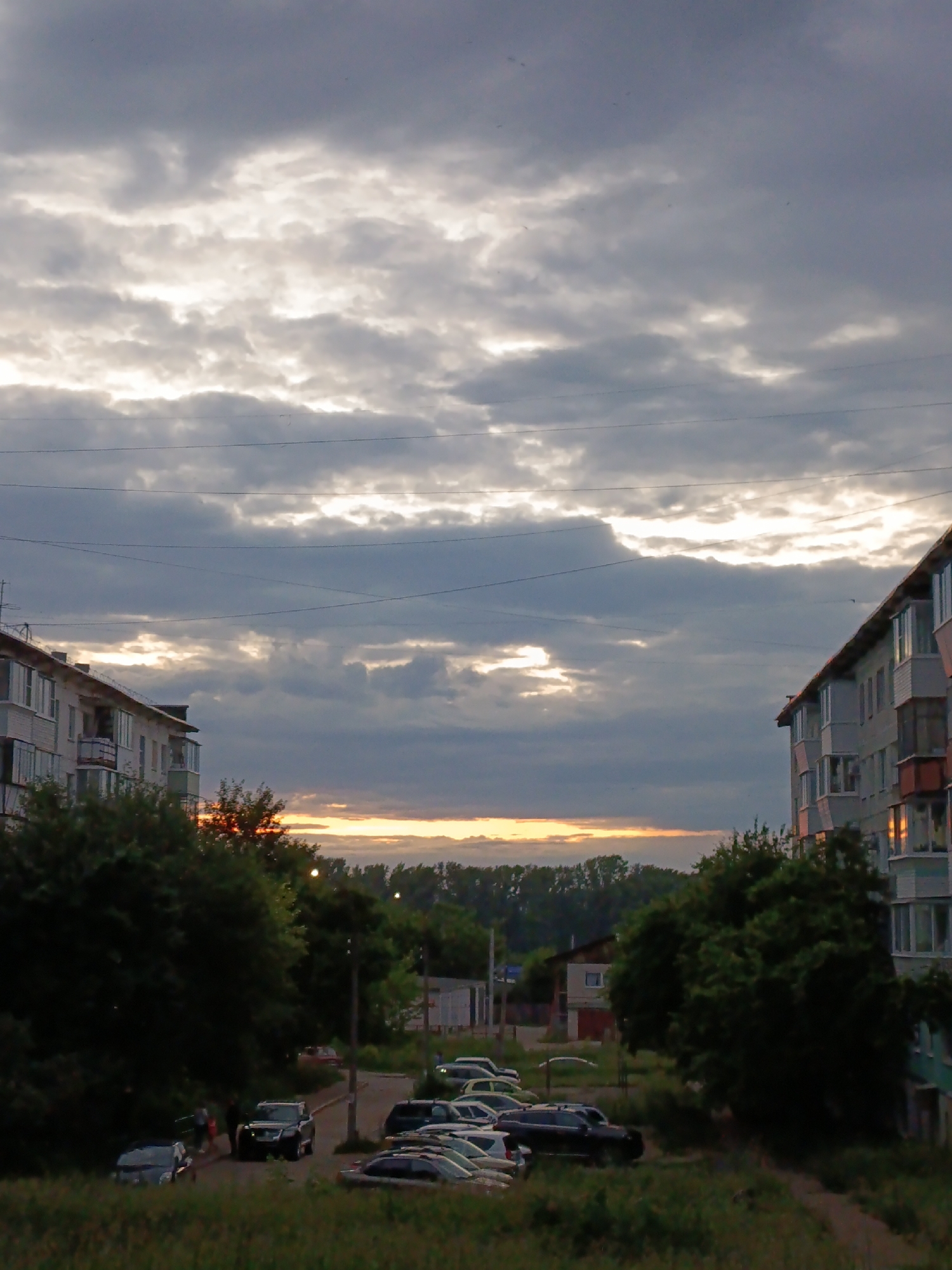 This is a daytime, eye-level, full shot of a residential street scene in Chaikovsky, Russia, at dusk. The sky is filled with dramatic, layered clouds, with breaks of sunlight illuminating the scene. Two apartment buildings, one on the left and one on the right, flank the street, which is lined with trees and parked cars. The building on the left is light-colored with visible windows and balconies. The building on the right is a multi-story structure with a darker facade, also featuring windows and balconies.
On the street, several cars are parked, including a black SUV in the foreground. A few individuals are visible near the cars on the left side of the street, appearing to be walking. The overall impression is of a quiet residential area at the end of the day, with the natural light creating a somewhat moody atmosphere. The environment appears to be a mix of urban residential structures and surrounding green vegetation.