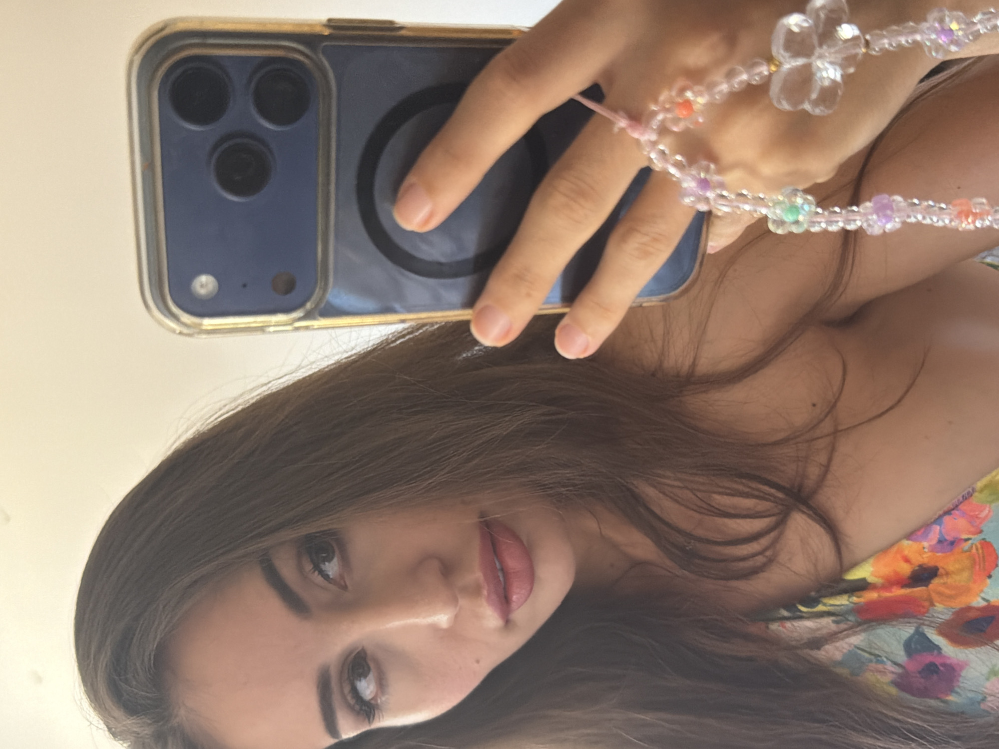A selfie taken indoors shows a woman holding a phone with a blue case and a beaded charm attached to her wrist. The woman has long brown hair and is wearing a floral-patterned top. The phone has a multi-lens camera system. The background appears to be a wall. There is no visible indication of the city of Tersefanou, Cyprus.
