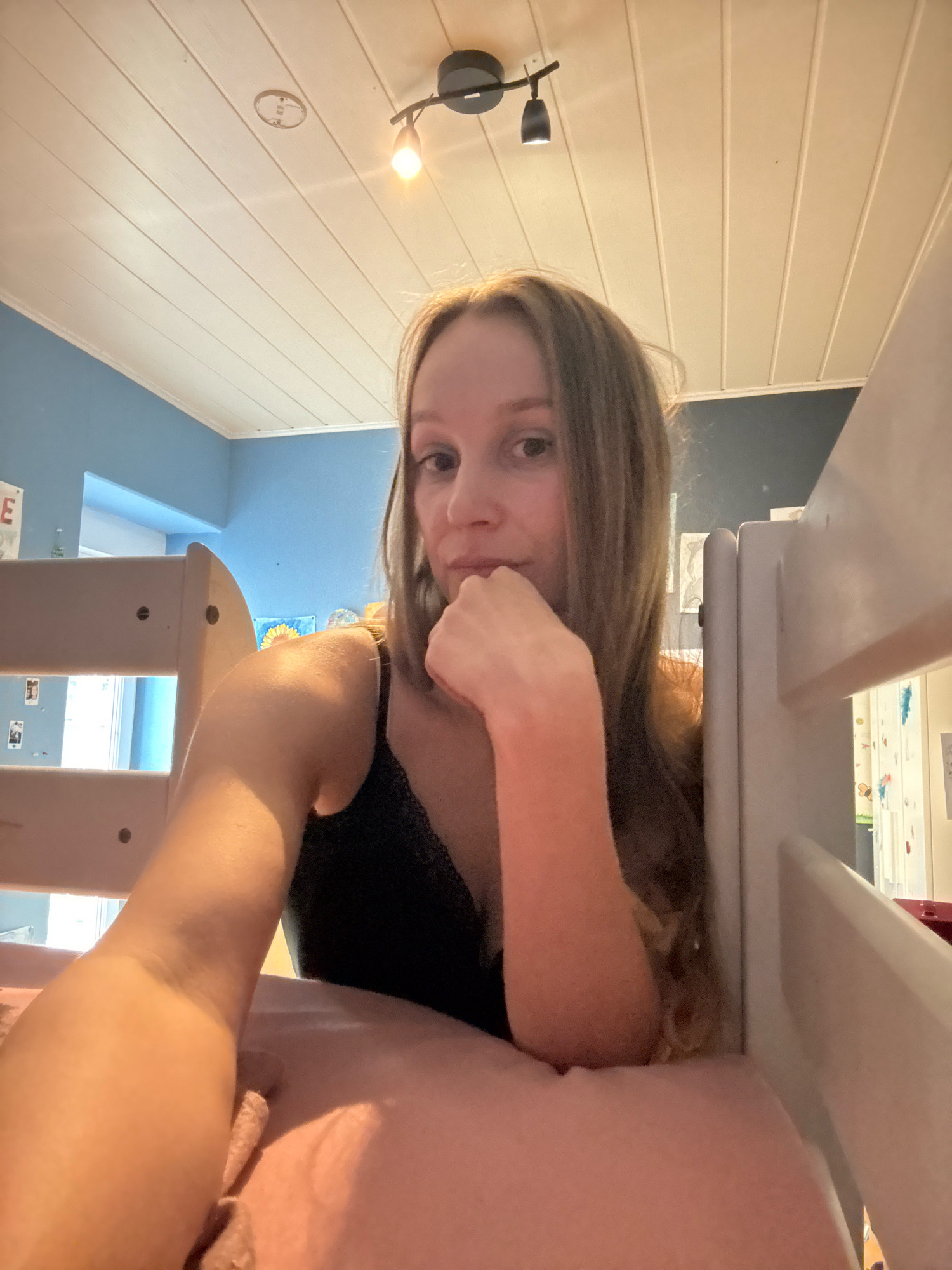 The image is a selfie taken from a low angle, featuring a young woman with long, blonde hair. She is wearing a black tank top with lace detailing. Her right arm is extended, and her hand is resting on her chin. The background shows a blue wall and a white, slatted ceiling with track lighting. To the right of the woman, a portion of a white bunk bed frame is visible, along with a pink pillow. The overall setting appears to be a bedroom. The specific location in Überherrn, Germany, is not discernible from the visual information provided.