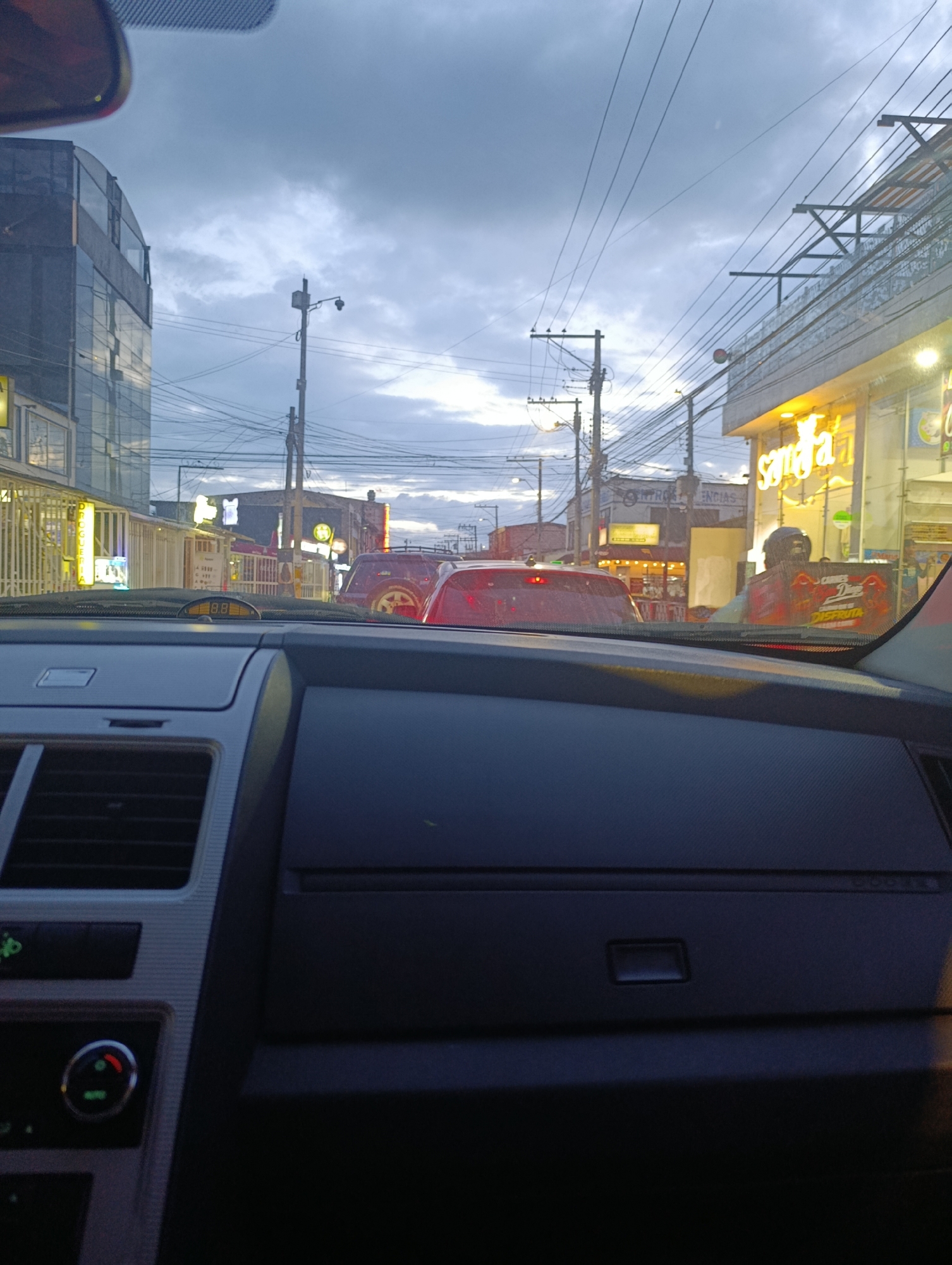The image presents a street view from inside a car, looking forward through the windshield at what appears to be a busy urban street during dusk or early evening. The sky is overcast with gray clouds, and artificial lights from buildings are already on.
In the foreground, the dark gray dashboard of the car is visible, featuring air vents and a climate control panel on the left with a visible "AUTO" button. A small, orange digital display on the dash shows the number "88." Part of a rearview mirror is blurred in the top left corner of the frame.
The midground depicts slow-moving traffic or a traffic jam, with several vehicles ahead. A red car is directly in front, and another red vehicle is further down the road. To the right, a person, likely a delivery driver, is on a motorcycle or scooter, carrying a large red delivery box. Text partially visible on the box includes "Carnes y..." and "DISFRUTA."
The background shows commercial buildings lining the street. On the left, a modern building with glass panels reflects the cloudy sky. Further along the street, a sign for "Drogueria" (pharmacy) is visible. On the right, a building with bright, illuminated yellow signs stands out, with the word "Santa" prominently displayed, suggesting a restaurant or business. Numerous utility poles and a dense network of overhead power lines crisscross the sky, characteristic of streets in Funza, Colombia. The scene captures a typical urban commute as day transitions to night.