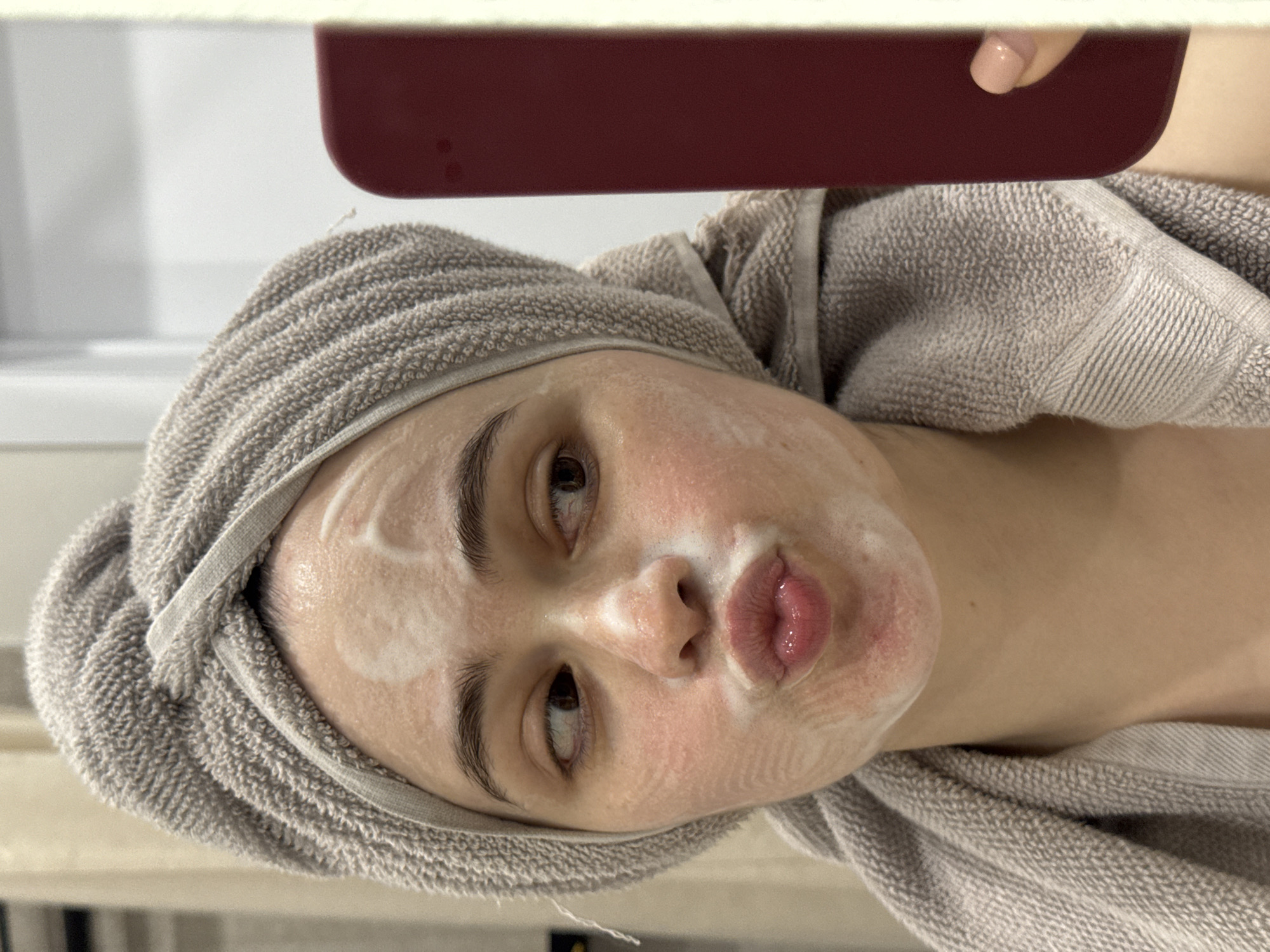A woman with fair skin and dark eyebrows is lying down. A light gray towel is wrapped around her head. Her face is covered in a white, creamy substance. She has brown eyes and her lips are pursed in a kissing expression. In the upper portion of the image, a corner of a red rectangular object is visible. The background is blurry and light-colored. There are no identifiable details that would indicate this image was taken in Izhevsk, Russia.