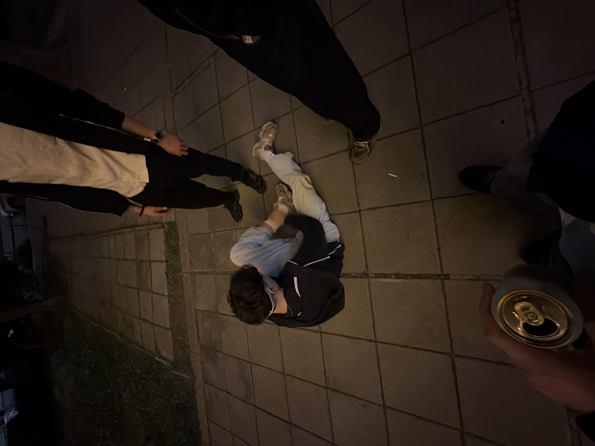 The image depicts a group of people standing and sitting on a tiled sidewalk at night. One person is sitting on the ground, wearing light-colored jeans and a dark jacket. Another person is holding an open aluminum can. The location appears to be an urban environment. Though not definitively confirmed, the tiled sidewalks are a feature in Sofia, Bulgaria.