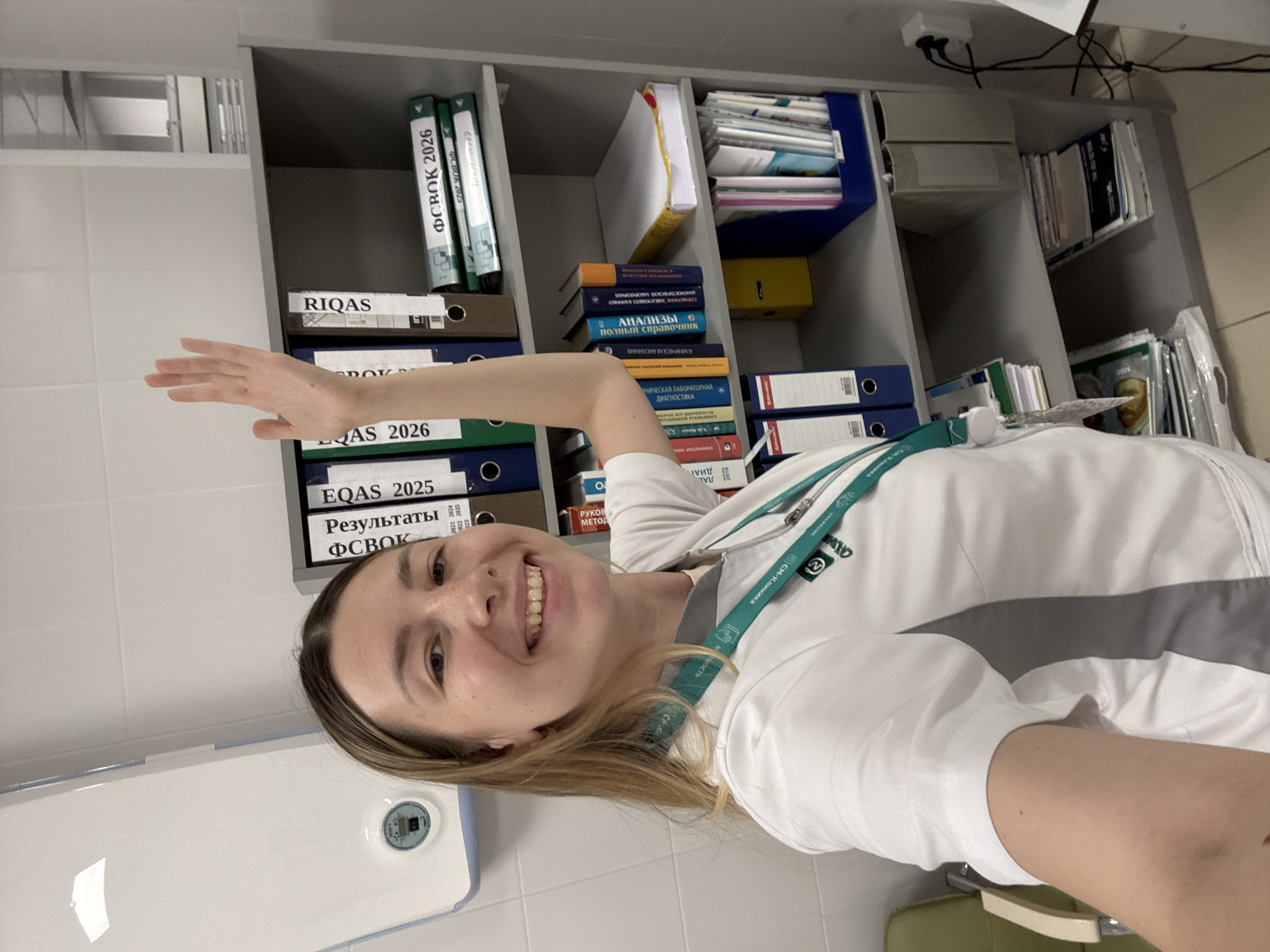 A woman with blonde hair, wearing a white lab coat with gray accents and a teal lanyard, stands in front of a grey metal shelving unit filled with binders and books. Her right arm is raised, with her palm open, as if waving or presenting something. The shelving unit is organized with various folders and publications, some of which have Cyrillic text visible, such as "EQAS 2025" and "ФСВОК 2026". To the left of the woman, a white medical device is mounted on a tiled wall. The background appears to be an indoor professional setting, possibly a laboratory or office. The image does not contain any specific information to determine its location within Moscow, Russia.
