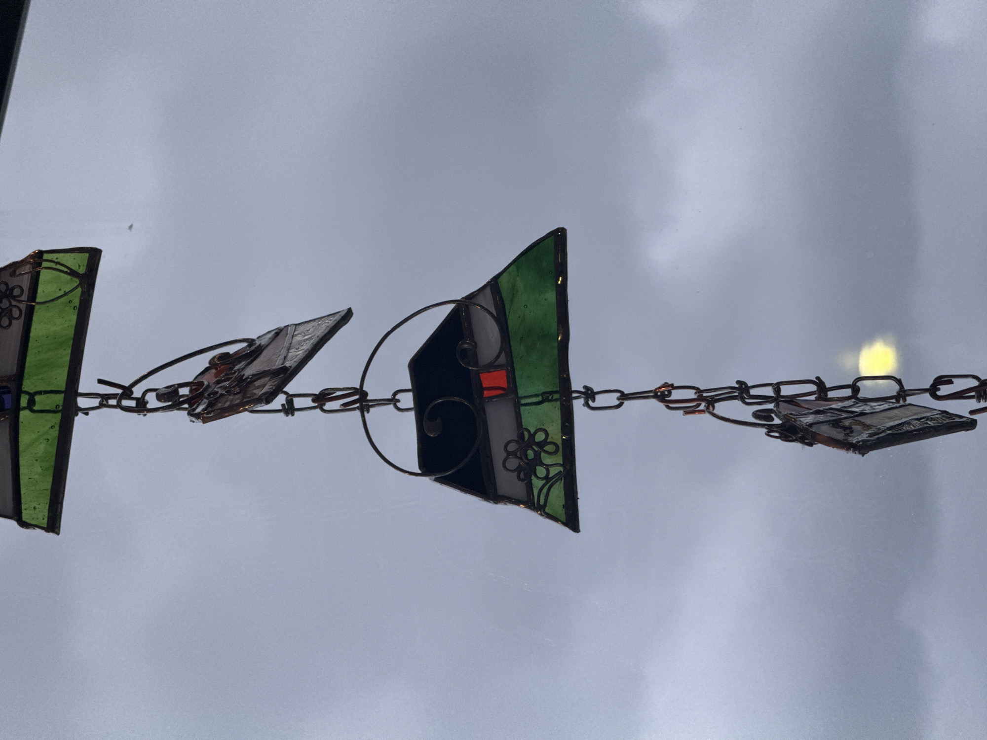 A close-up shows a chain of stained glass pieces suspended against a backdrop of overcast sky. The pieces are roughly rectangular and feature various colors and patterns, including green, gray, black, and red. The chain is made of dark metal links, and the pieces are attached to it via metal loops. The location and other city details are unknown.