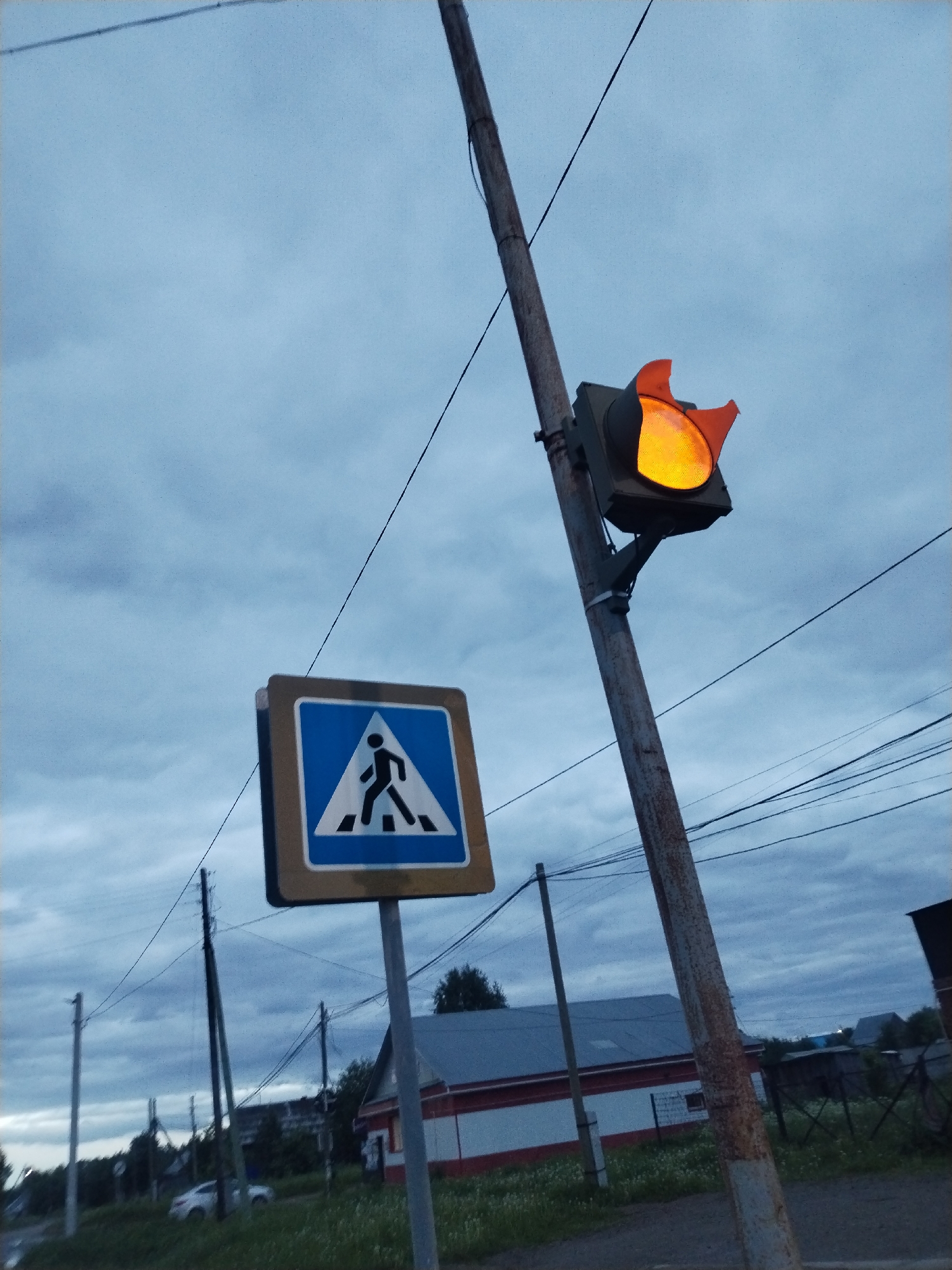 The image depicts a street scene in Сосново, Russia, under a cloudy sky. A pedestrian crossing sign, displaying a blue square with a white silhouette of a person walking on zebra stripes, stands next to a rusted pole. Mounted on the pole is an illuminated amber traffic light with damaged visors. Overhead power lines intersect the scene. In the background, there is a building with a gray roof, white walls, and red trim. Vegetation and a car are visible on the side of the road. Other utility poles appear further in the distance.