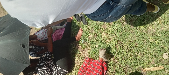 The scene is a grassy outdoor area in Kitengela, Kenya, located at coordinates -1.473 latitude and 36.957 longitude. Two women and a young child are sitting on the grass. One woman is wearing a black and white striped shirt and black pants, while the other woman is wearing a black shirt and black pants. The child is wearing a white shirt and denim shorts and is being held by the second woman. The first woman is holding a black umbrella over the group. The women are facing the camera, while the child is facing away. Another person, whose clothing is partially visible, is standing behind the group. They are wearing a white shirt and blue denim pants. The person is looking at the camera. The ground is covered in grass, and there are some small rocks scattered around. The sun is shining and casting shadows on the grass.