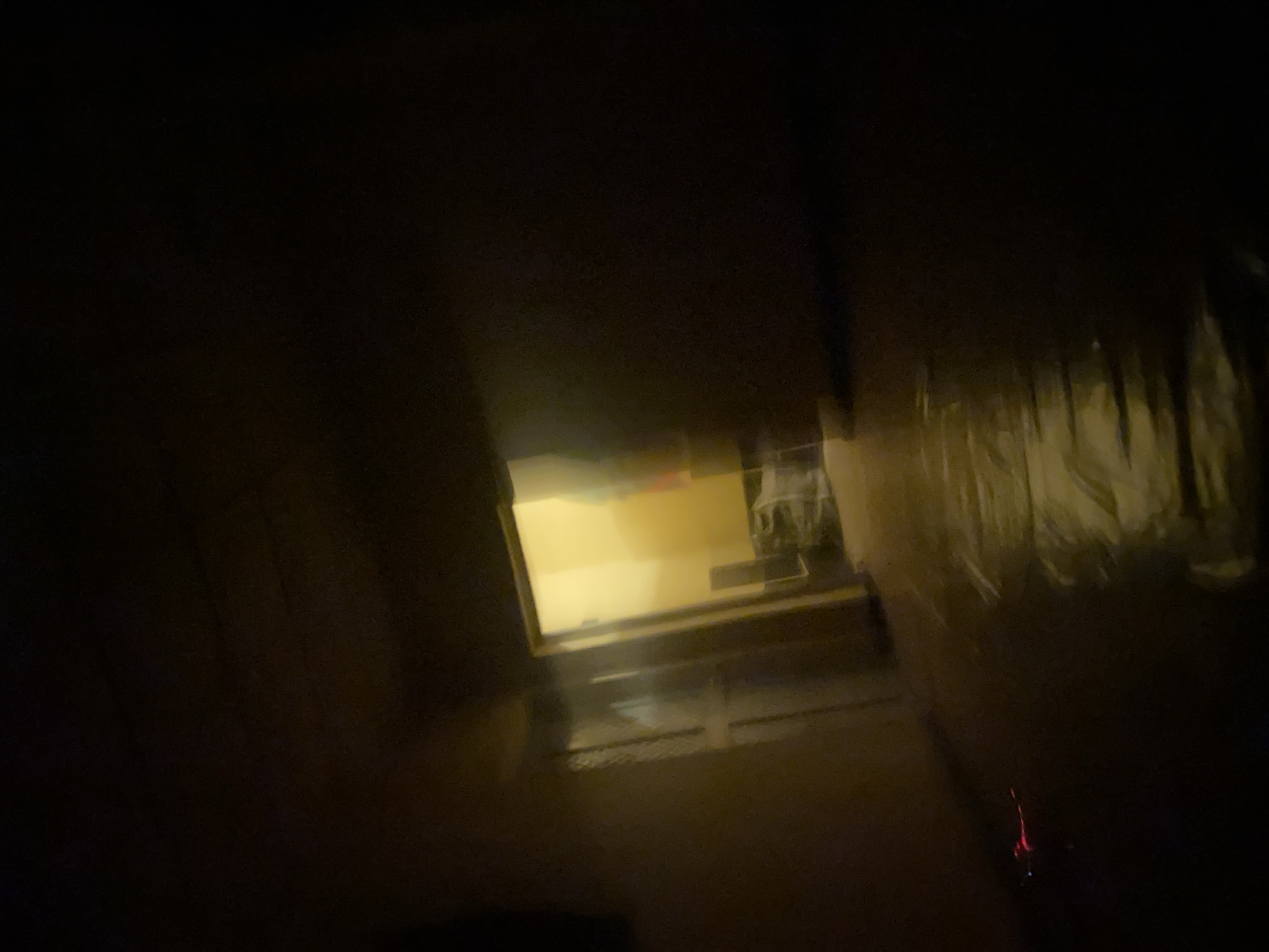 The image shows a dimly lit hallway with a door open at the end, revealing a yellow-lit room. The hallway walls are textured and appear to be painted. The floor is covered with a reflective material, possibly plastic sheeting, which catches the light. In the foreground, to the left, a small, brightly lit red object is visible on the floor. The overall atmosphere is dark and somewhat eerie, with limited visibility. The specific location in Barikin Jirga Summa, Nigeria cannot be determined from the visual information provided in the image.