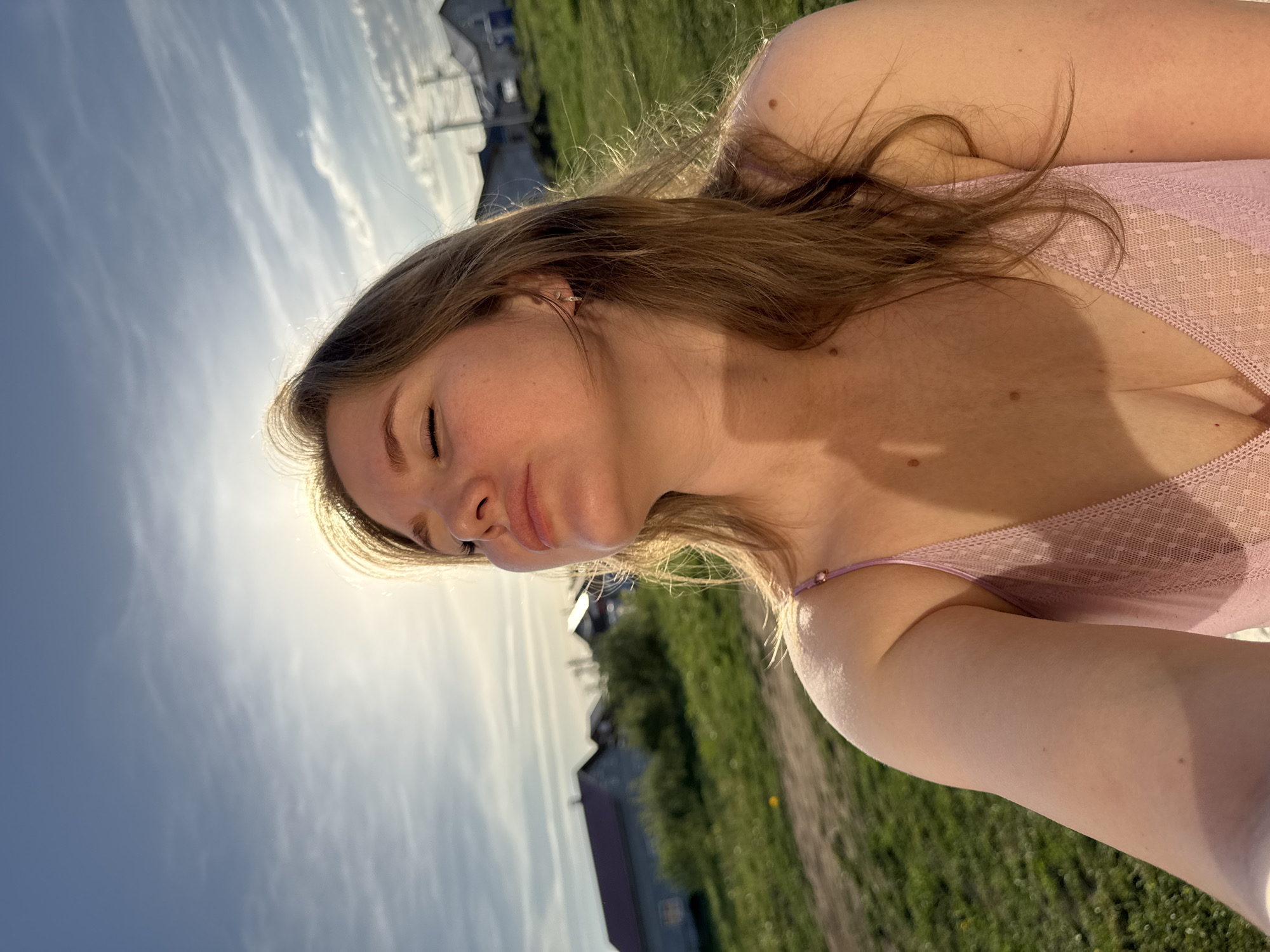 The image depicts a woman outdoors. She has fair skin and light brown hair. She is wearing a pink top with lace trim. The background shows a grassy field and buildings, consistent with a rural or suburban setting. The sky is visible in the background with a few clouds and the sun positioned above the woman's head, which is in the city of Ферма, Russia.