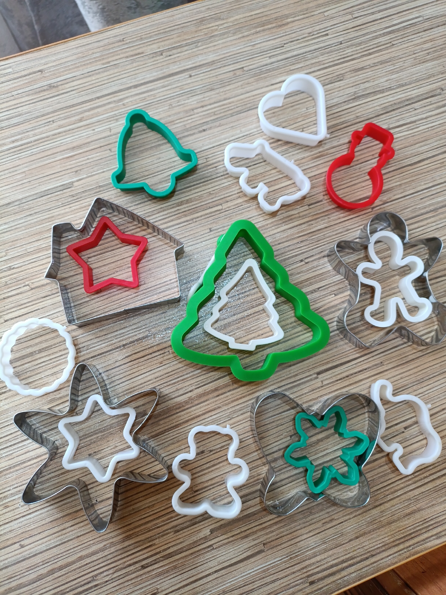 The image displays a collection of 12 distinct cookie cutters arranged on a light-colored wooden surface, exhibiting prominent horizontal wood grain. The objects are scattered without a specific pattern.
The cookie cutters vary in material and color:
* Six are silver-colored metal (likely stainless steel).
* Six are plastic, consisting of three primary colors: white (5 objects), green (2 objects), and red (2 objects).
Specific cookie cutters and their arrangements, from top-left to bottom-right:
1. A green plastic bell.
2. A white plastic heart.
3. A white plastic mitten or glove.
4. A red plastic snowman.
5. A silver metal house-shaped cutter, containing a nested red plastic star.
6. A white plastic wavy circular ring.
7. A green plastic Christmas tree cutter, containing a nested white plastic Christmas tree.
8. A silver metal gingerbread man cutter, containing a nested white plastic gingerbread man.
9. A silver metal star cutter, containing a nested white plastic star.
10. A small white plastic gingerbread man.
11. A silver metal butterfly cutter, containing a nested green plastic multi-pointed star or snowflake.
12. A white plastic abstract shape, resembling a stylized figure.
The wooden surface occupies the majority of the frame, with its grain running generally from upper left to lower right. The upper left corner shows a slight blur, indicating a minimal background element or the edge of the surface. No individuals are present in the image. No actions are depicted. The environment is an indoor setting, likely a tabletop. The image provides no visual information to determine its geographical location in the city of Chaikovsky, Russia, or any other specific place.
