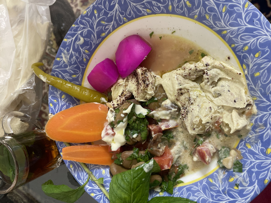 The image displays a close-up, top-down view of a meal presented on a blue and white patterned ceramic plate with a yellow rim, situated within an indoor setting in the city of Al-Qanawat Municipality, Syria. The plate contains Foul Medames (fava beans) mixed with chopped tomatoes, green herbs, and a creamy white sauce, with visible olive oil. Adjacent to this, a portion of light-colored mashed chickpeas or hummus is topped with sumac and olive oil. Accompanying items on the plate include two distinct, oblong slices of cooked orange carrot, one long green pickled pepper, and two bright pink pickled turnip pieces. Fresh green mint sprigs serve as a garnish on the left side of the plate. To the upper left, a clear glass cup with a handle contains a dark amber liquid, consistent with tea, featuring green mint leaves floating on its surface. A crumpled transparent plastic bag is partially visible in the background, behind the plate. The entire arrangement rests on a subtly patterned surface, possibly a textile or mat.