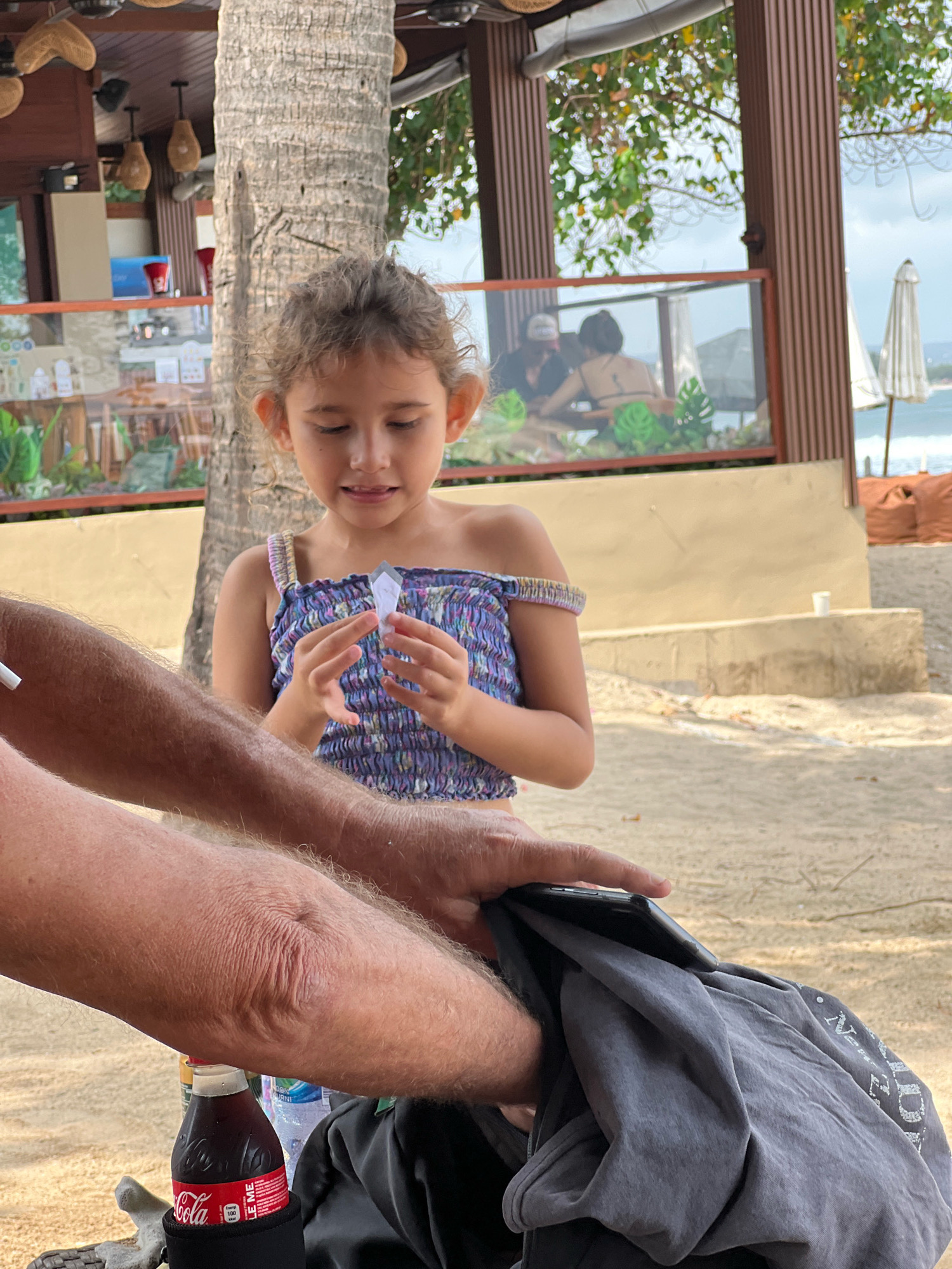 A young girl is standing on a sandy surface, holding a small, clear object in her hands. She is looking down at it with a curious expression. She is wearing a purple and blue patterned top. In the foreground, a person's arms and legs are visible. One arm is bent, and the other is resting on a gray garment that is partially covering a phone. A bottle of Coca-Cola is visible next to the person's leg. The background shows a beach with some umbrellas and the ocean in the distance. There is a wooden structure and a palm tree behind the girl. The scene appears to be outdoors, possibly at a beachside cafe or restaurant in Kuta, Indonesia.