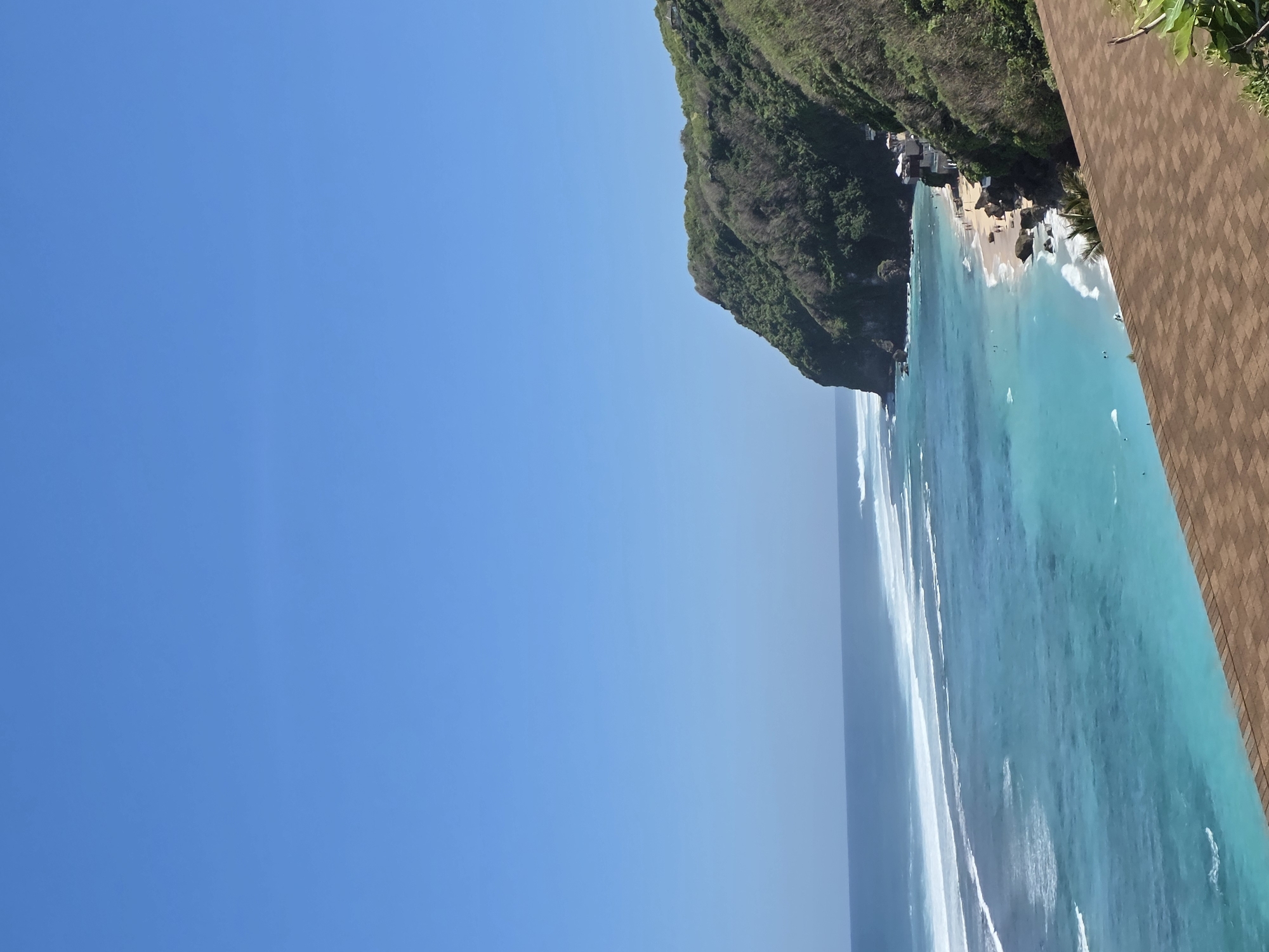 The image captures a coastal scene in Kuta Selatan, Indonesia, featuring a vibrant turquoise ocean with visible waves breaking along the shore. A verdant cliff covered in dense vegetation rises from the ocean, providing a dramatic contrast to the blue sky and water. On the beach, small structures or buildings are clustered near the base of the cliff, suggesting a small settlement or resort. A section of a brown tiled structure is visible in the corner of the image, likely a part of a building's roof or wall. The sky is clear and blue, indicating a sunny day.