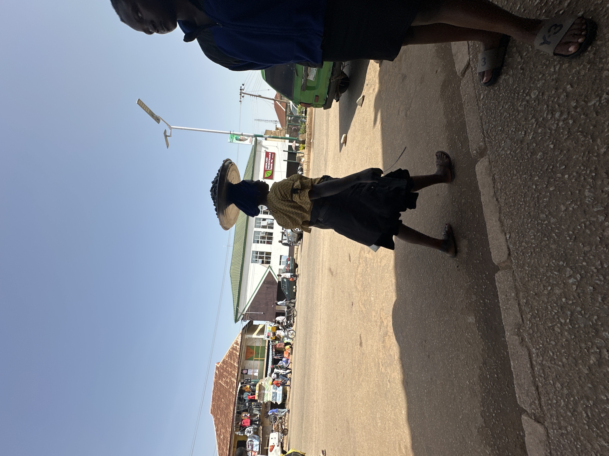 A wide shot captures a street scene in Bukuru, Nigeria, bathed in the bright light of a clear day. In the foreground, the legs and feet of two individuals are visible. To the left, a person walks in dark sandals, their shadow cast on the asphalt road. To the right, another person stands wearing Y-3 slides.
Further back, a woman, viewed from the side, walks with a large platter of dark items balanced on her head. She wears a patterned yellow top with ruffled sleeves and a dark skirt. She is walking on the road, which is paved with asphalt and shows some wear.
The background reveals a street with several buildings and vehicles. On the left, a two-story building with a reddish-brown tiled roof houses shops displaying clothing on its facade. Across the street, a white building with a green roof has windows and a sign that reads "LAGOS AND HIGHLAND MICROFINANCE BANK PLC." A green van is parked in front of this building. Various other vehicles, including a motorcycle, are also present on the street. Power lines and a modern streetlight are visible against the blue sky. The overall environment suggests a busy urban setting.