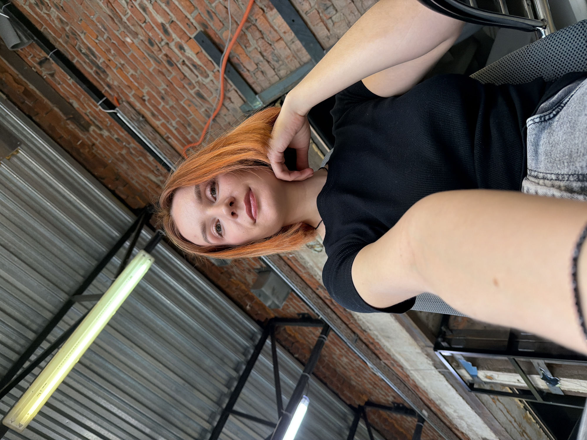 A medium-angle shot features a woman with light skin and reddish-orange hair. She is wearing a black top and what appears to be light-wash denim shorts. Her right hand is near her face, and her left hand is at her side. The location shows exposed overhead industrial elements and brickwork suggesting the city of Chaikovsky, Russia. In the background, there are metal beams and what appears to be a fluorescent light fixture. There is also an orange cable running along the brick wall. The angle of the shot indicates that the photo was likely taken from a low perspective, looking up at the woman.