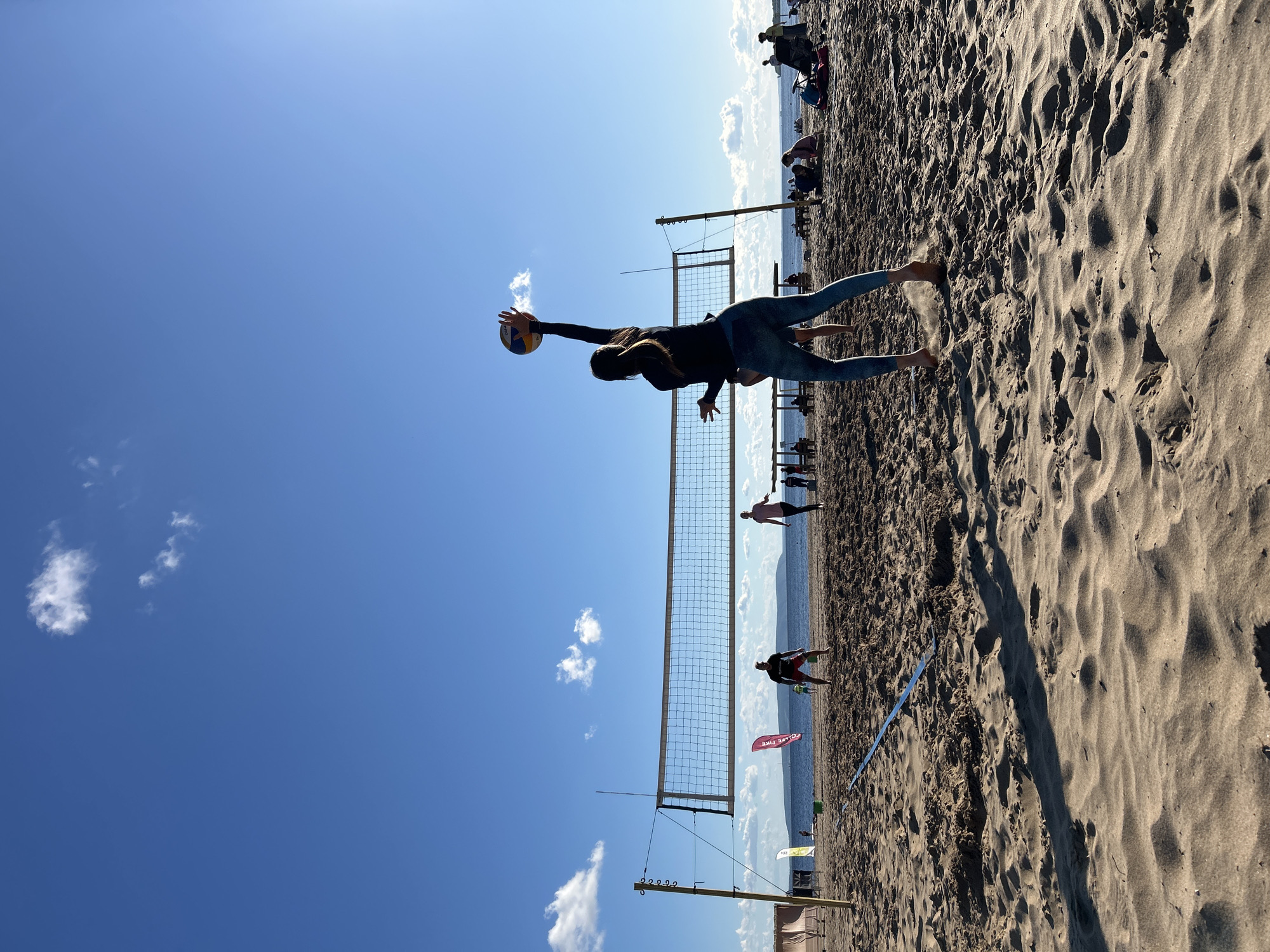 The image depicts a sunny day at a sandy beach, likely in Tolyatti, Russia, given the context. The sky is a clear, vibrant blue with a few scattered white clouds. A volleyball net is set up on the sand, indicating a beach volleyball game is in progress or has recently occurred.
The main subject is a person, silhouetted against the bright sky, mid-air as they dive to hit a volleyball. They are wearing a dark long-sleeved shirt and light-colored leggings. Their body is extended, one arm reaching out to make contact with the ball, which is also visible.
In the background, other people are present on the beach, some playing or spectating near the net, while others are further back, possibly relaxing or walking along the shoreline. The sea or a large body of water is visible in the distance, with a hazy horizon. The sand is textured with ripples and shadows, suggesting the sun is high in the sky, likely late morning or early afternoon.
There is a red flag planted in the sand with some white text on it, but it is partially obscured and illegible. Another smaller white flag is visible further down the beach. The overall mood is active and energetic, capturing a moment of athletic play on a beautiful, clear day.