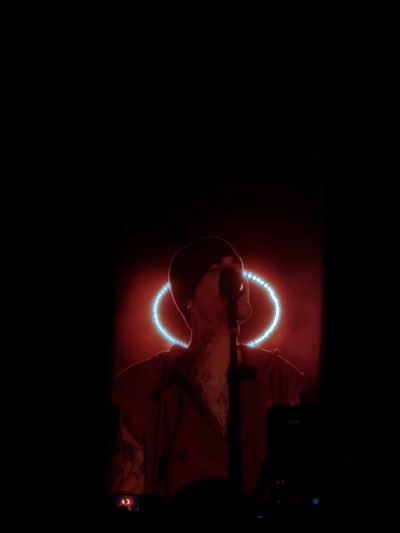A performer is on stage at a venue in Indio, United States. The person is wearing a beanie, and a microphone stand is in front of them. A blue circle behind the performer illuminates the scene.