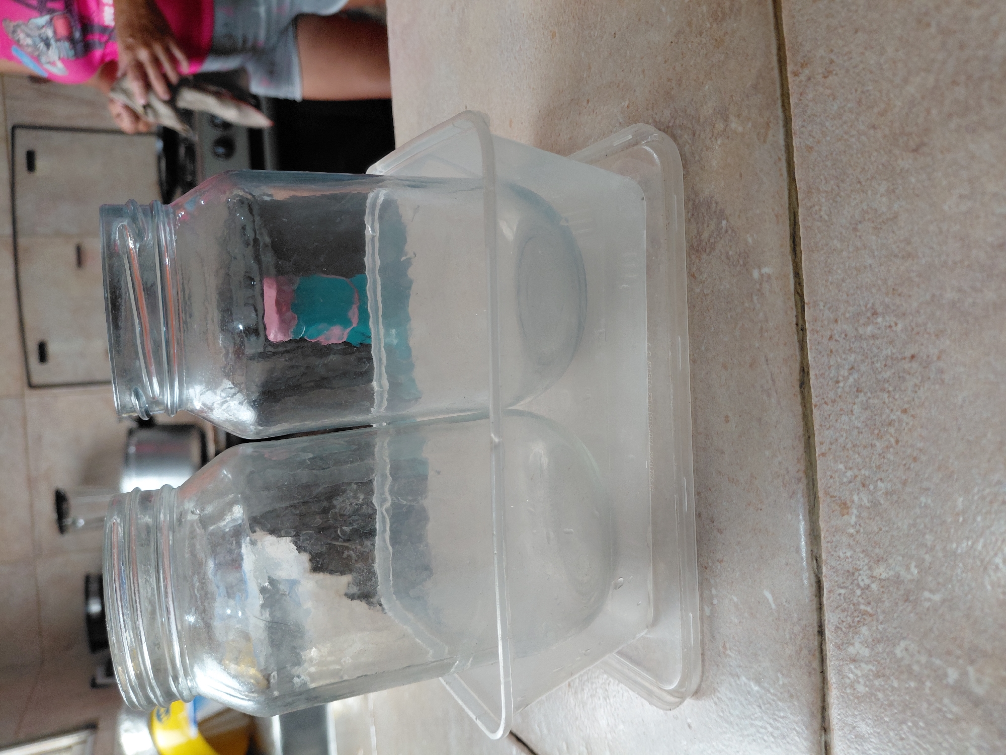 The image displays a scene inside a kitchen, rotated 90 degrees clockwise. In the foreground, two clear, empty glass jars with threaded necks are placed upside down within a rectangular, clear plastic container. The plastic container appears to be functioning as a drying rack or holder for the jars. Faint, molded text is visible along the rim of the plastic container, though it is largely illegible. The items rest on a light brown or beige tiled countertop, characterized by distinct dark grout lines.
In the blurred background, a person is partially visible, dressed in a vibrant pink t-shirt and grey shorts. Their hands are occupied with an object, possibly a utensil or food item, over a dark kitchen appliance that resembles a stove or oven, with some controls discernible. Another kitchen fixture, possibly a sink or cabinetry, can also be seen in the background. The lighting is bright, suggesting it is daytime. The overall scene depicts a casual, everyday kitchen activity, likely involving food preparation or clean-up. There are no discernible emotions from the partial view of the person.