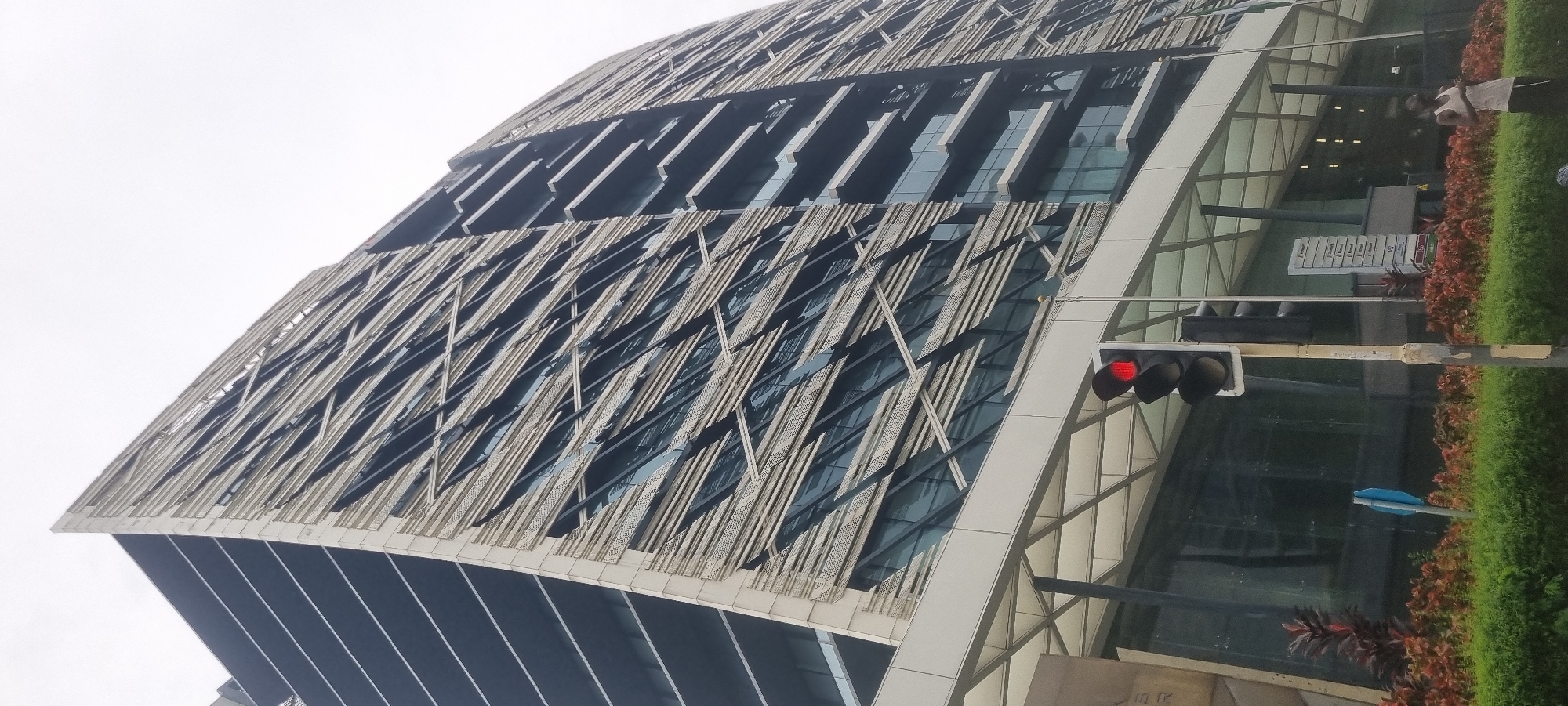 The image shows a low-angle view of a modern, tall office building in Ikoyi, Nigeria. The building has a distinctive facade with horizontal white fins that create a textured, angular pattern over dark glass windows. The top of the building is sharply angled and protrudes outwards. In the foreground, a traffic light with a red light illuminated is visible, along with a street sign. A person wearing a white tank top and dark shorts stands to the right of the traffic light. Lush green bushes and red-leafed plants line the area in front of the building. The sky is overcast, suggesting a cloudy day. The overall scene depicts an urban environment with a focus on the architecture of the building and its immediate surroundings.