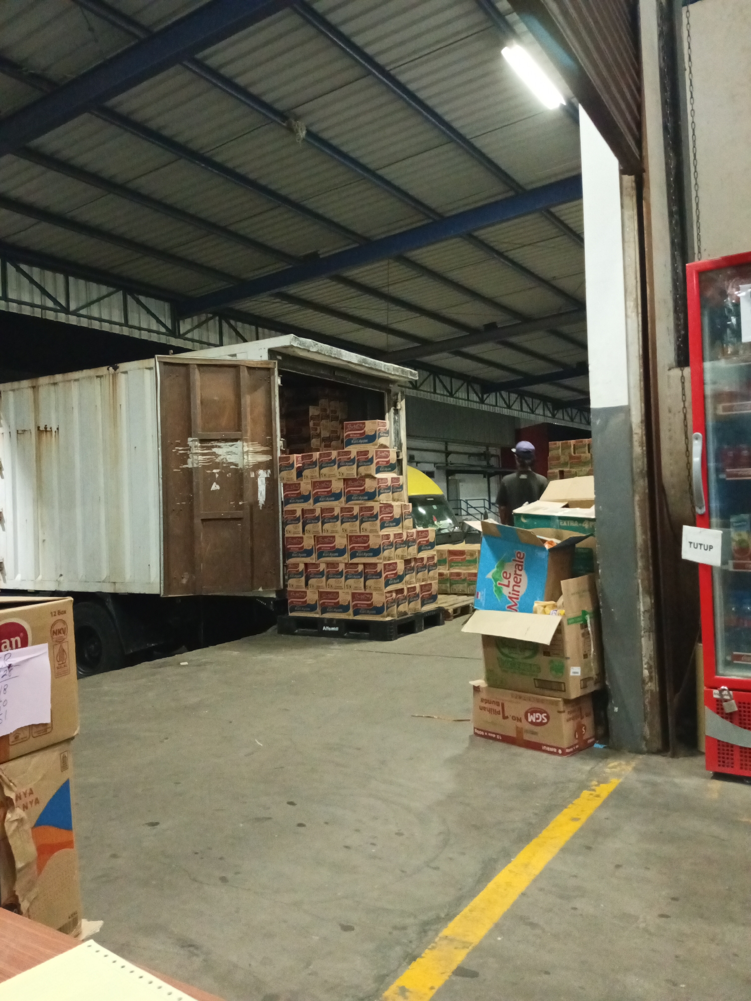 The image depicts the interior of a warehouse in Makassar, Indonesia, likely a distribution center for consumer goods. The scene is dimly lit, with artificial lights from the warehouse ceiling illuminating the area. In the foreground, a concrete floor is visible with a prominent yellow line marking a path or boundary. Several cardboard boxes are stacked, some appearing to contain products like instant noodles and powdered milk.
To the left, a large white cargo truck with its rear doors open is parked. The back of the truck is filled with stacks of identically branded boxes, suggesting a recent delivery or pending shipment. A portion of the truck's side is also visible, showing signs of wear and tear.
In the middle ground, a forklift is partially visible, hinting at ongoing logistical operations. Beyond the truck, a yellow vehicle is parked, and a person wearing a blue cap and dark shirt is standing near it, facing away from the camera.
To the right, a tall, red commercial refrigerator or cooler is positioned against a wall, with a white "TUTUP" (Closed) sign attached to it. The cooler appears to be stocked with beverages. The overall environment suggests a functional, albeit somewhat cluttered, industrial space dedicated to the storage and movement of goods.