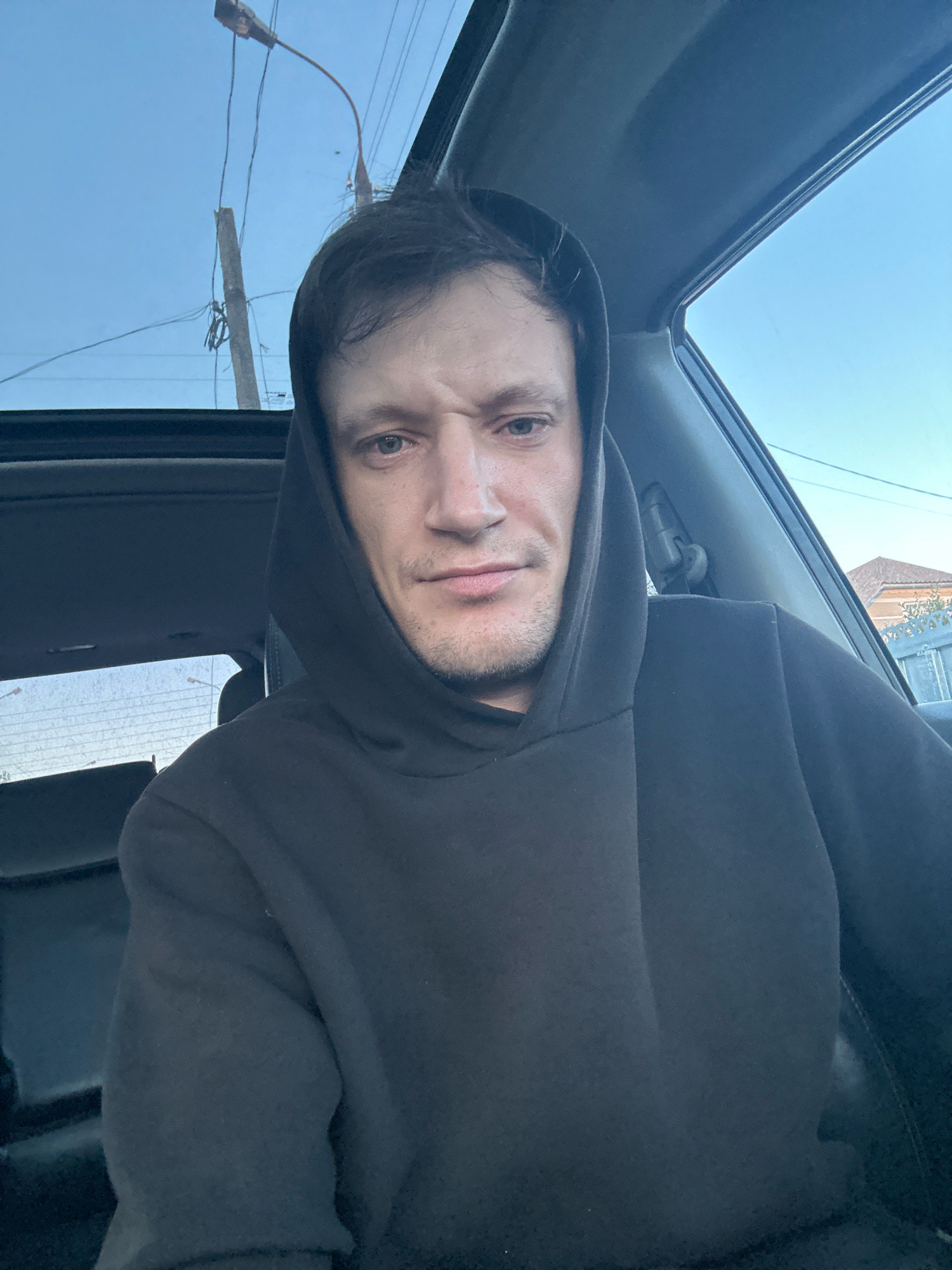 The image is a close-up selfie of a man inside a car. He is wearing a dark hooded sweatshirt and looking directly at the camera. The man has light-colored eyes and stubble on his face. The interior of the car, including the black seats and a sunroof, is visible. Outside the car, the sky is blue, with power lines and a street light visible. In the background, a house and a fence can be seen. The photo was taken in Makiv, Ukraine.