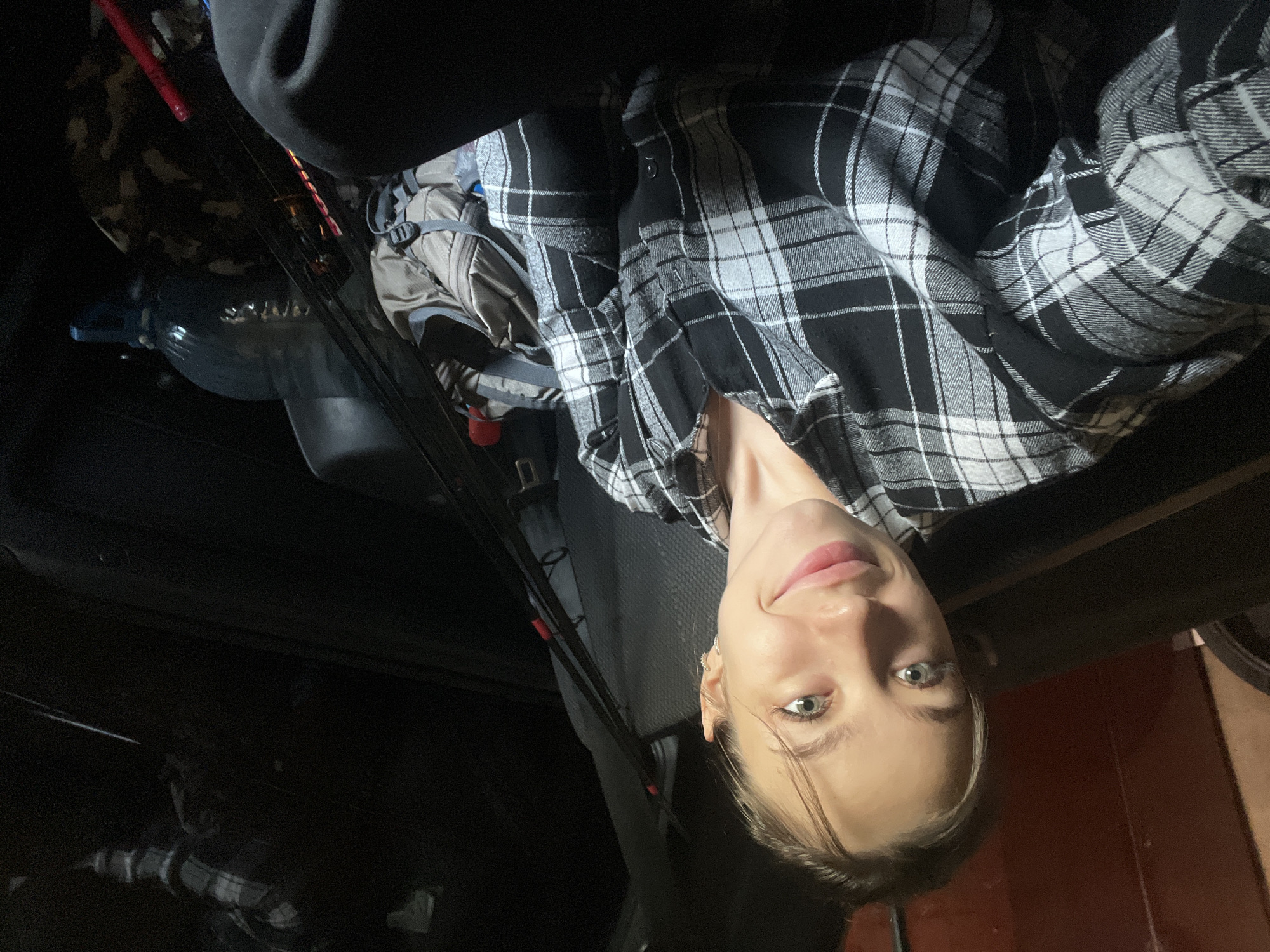 A young woman with light skin and light brown hair pulled back is looking at the camera, smiling. She is wearing a black and white plaid shirt. She appears to be in the back of a vehicle. To her right, there are fishing rods, a backpack, and a large blue water bottle. The interior of the vehicle is dark, with some reflections visible. The scene suggests preparation for an outdoor activity or travel.