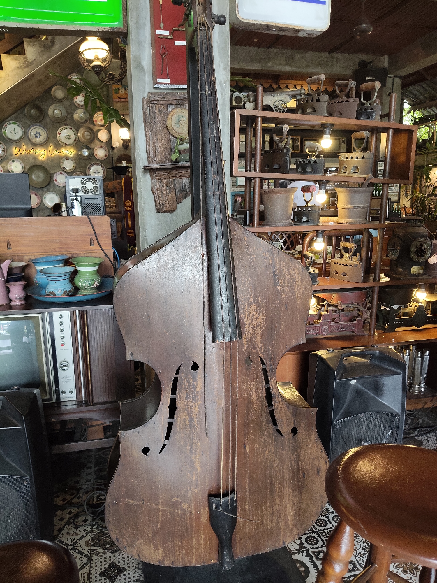 This image depicts a dimly lit, eclectic interior space that appears to be a cafe or restaurant in Medan, Indonesia. The focal point is a large, weathered wooden double bass standing upright in the foreground. To the left of the double bass is an old television set and a shelf filled with various vases. Behind the double bass, a wooden shelving unit is laden with an assortment of antique items, including several cast-iron irons and other metal tools. To the right of the double bass, a black speaker sits on the floor, and a wooden stool is partially visible. In the background, shelves are filled with more vintage objects, and a staircase with plates displayed on the wall is visible on the far left. The overall atmosphere is one of rustic charm and nostalgic decor.