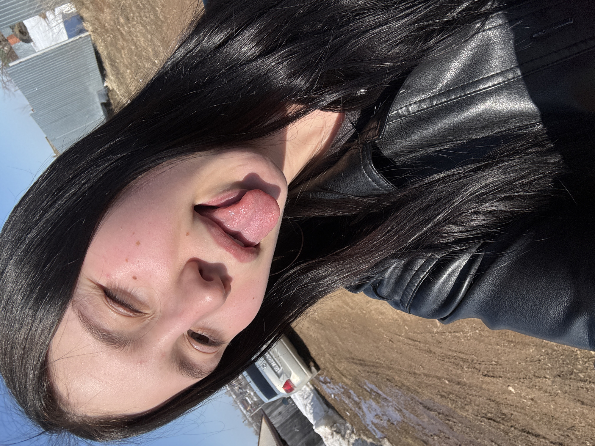 A rotated close-up shot captures a young woman with long, dark hair wearing a black leather jacket. She is sticking out her tongue and winking. Her skin is fair, with some freckles visible on her forehead and cheek. The background includes a partially visible white car and what appears to be a field or a barren landscape, with some snow patches still present. In the far background, there is a glimpse of a building with a metallic roof. The car's license plate is partially visible, with the letters "WX374" identifiable. The overall setting suggests an outdoor location, likely in the city of Фоки, Russia, given the landscape and architecture.