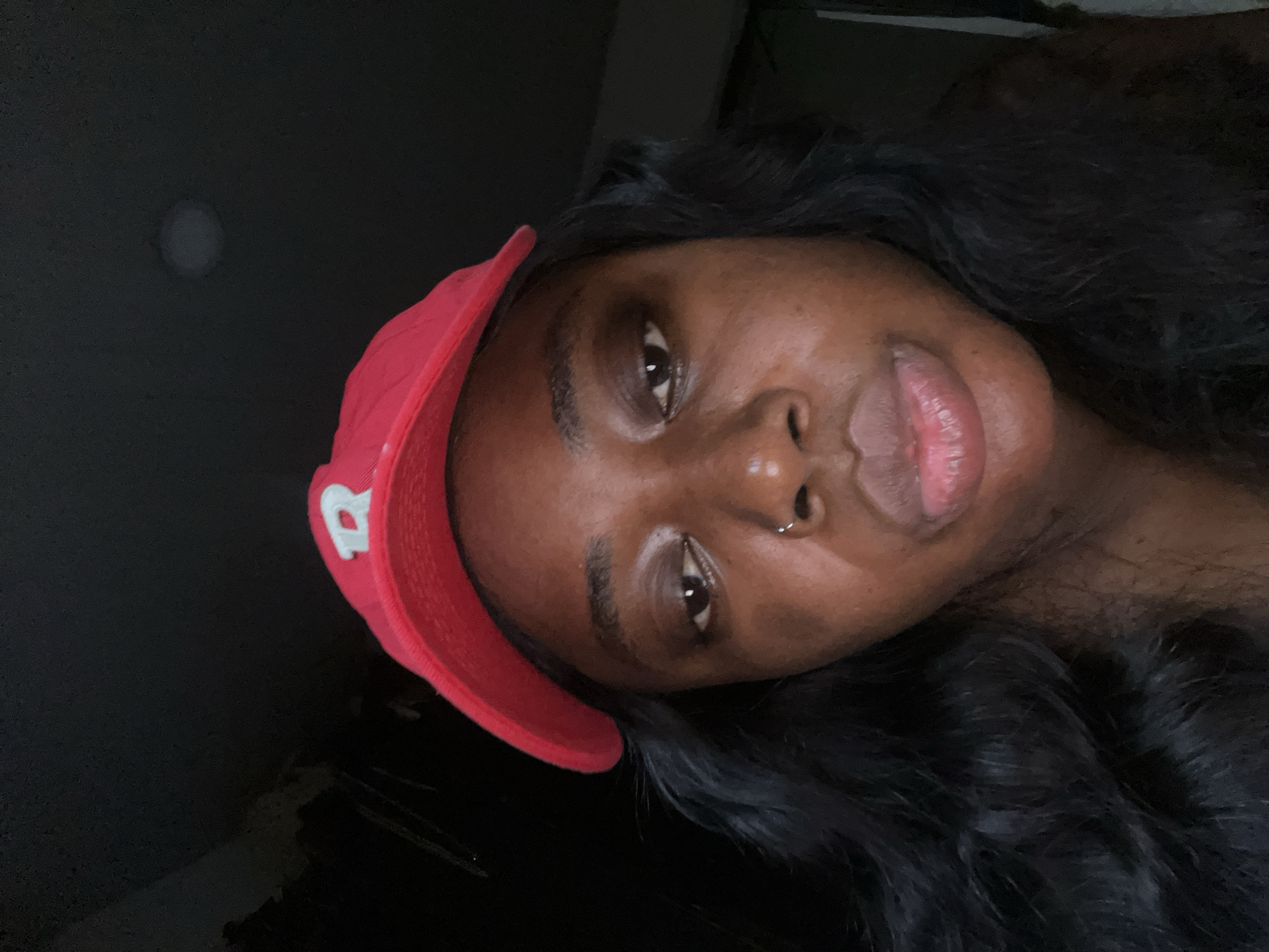 A young woman with dark skin and long, dark hair wears a red baseball cap with a white embroidered "R" on it. She is wearing a nose ring. Her lips are slightly parted. The lighting in the image is dim, with a light source above and to the right casting a slight glow on her face. The background is dark and out of focus. There is no discernible location in Abuja, Nigeria.