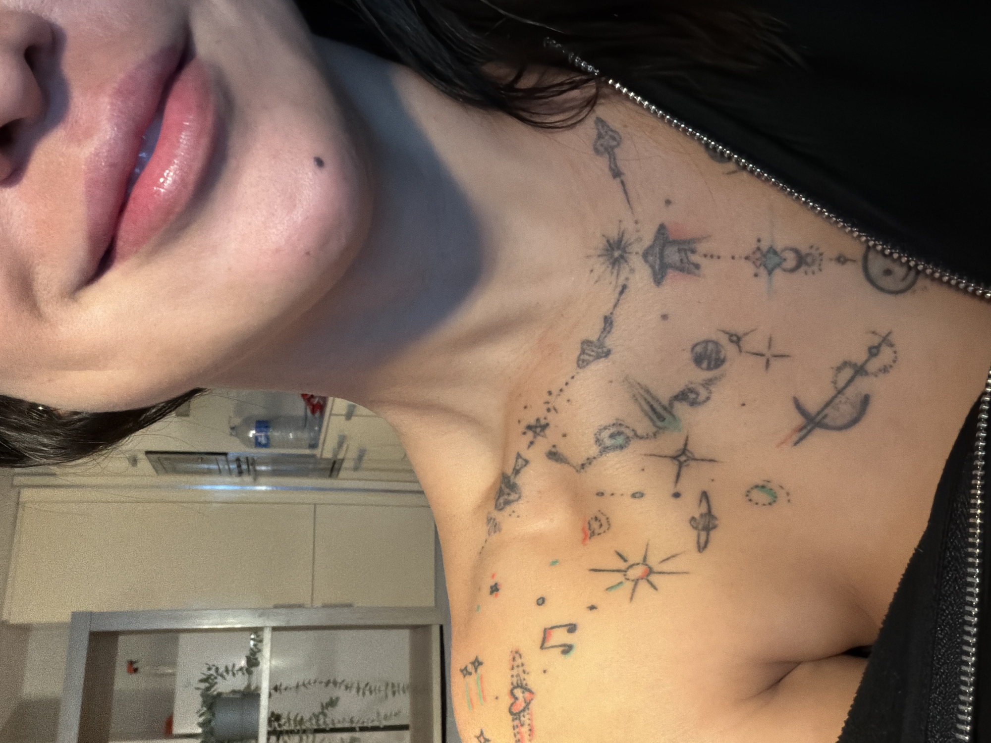 The image shows a close-up view of a person's face and upper chest. The person has fair skin, dark hair, and full lips with a glossy finish. A mole is visible on the side of the chin. The person has numerous tattoos on their chest and collarbone area, including stars, constellations, a moon, an anchor, and other symbols. The person is wearing a black zippered jacket. The background appears to be a kitchen, with beige cabinets, shelving, and a plant visible. There is no definitive evidence to confirm the location is L'hospitalet De Llobregat, Spain.