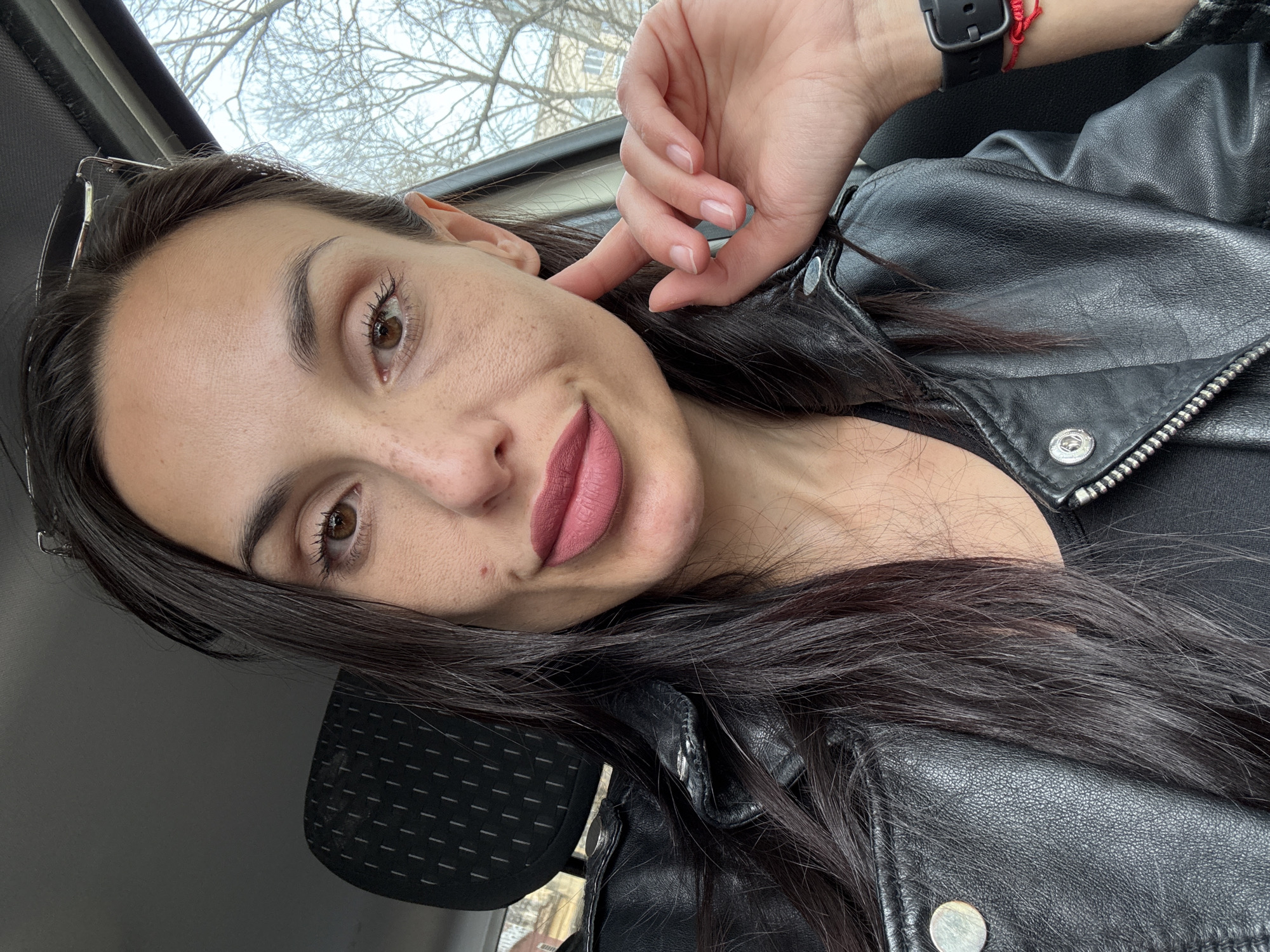A woman with dark hair and brown eyes is shown in a close-up, angled selfie. She wears a black leather jacket and a black wristwatch with a red bracelet on her left wrist. Her hand is near her face, touching her cheek. The background shows the interior of a car with a headrest and a window with trees visible outside. The location is Pleven, Bulgaria.