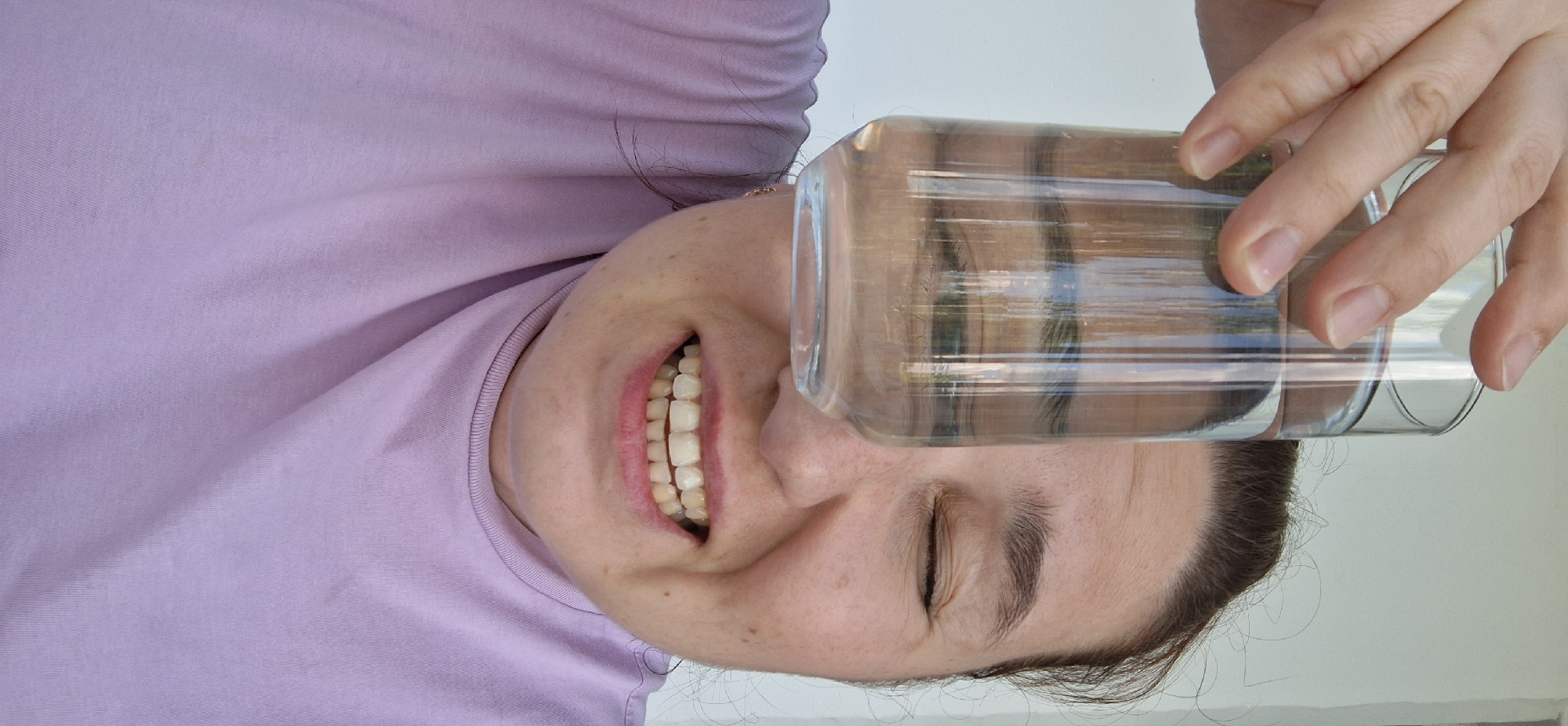 A woman with fair skin and dark hair is holding a clear glass with liquid in front of her face, distorting her closed eye. She is smiling, revealing her teeth. She wears a lavender t-shirt. The background is a plain, light wall. The location cannot be determined from the image.