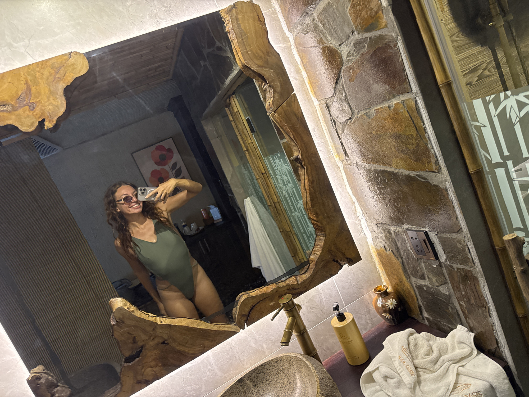A woman in a green one-piece swimsuit and sunglasses smiles at her reflection in a large, irregularly shaped mirror with a wooden frame. She is taking a selfie with a phone. The bathroom features stone walls, bamboo accents, and a wooden countertop. Visible objects include a faucet, a soap dispenser, a small decorative vase with flowers, and a folded white towel with embroidery. The room appears to be a high-end spa or hotel bathroom, possibly located in Nam Hoa Lư Ward, Vietnam, given the apparent architectural style and bamboo decor.
