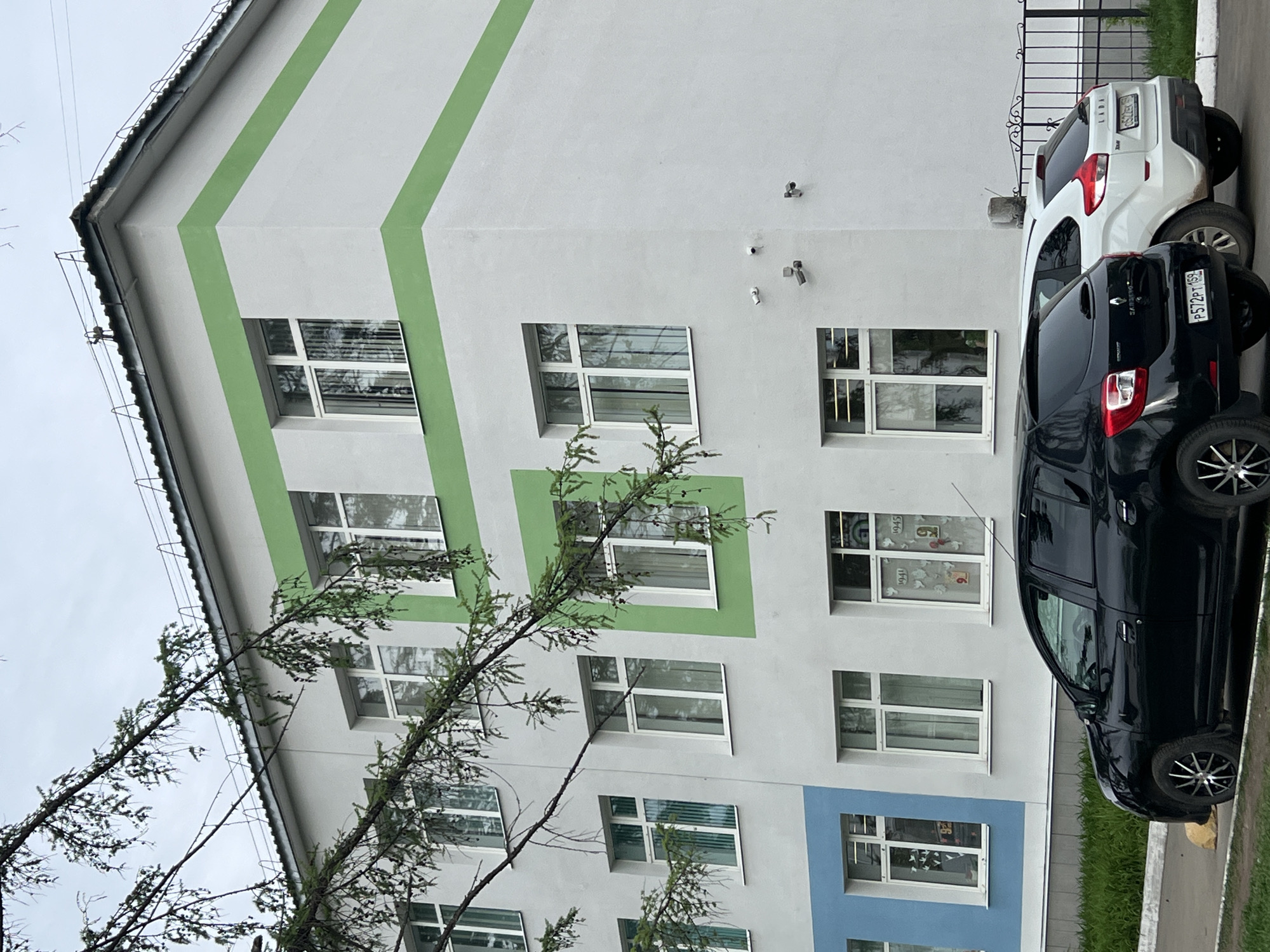 The image shows the facade of a building, likely in Chaikovsky, Russia. The building is painted white with green accents around the windows, except for one blue accent. A tree partially obscures the view of the building's lower portion. Two cars, one white and one black, are parked alongside the building. The license plate of the black car is partially visible, showing "P572PT" and the Russian flag. Surveillance cameras are visible on the building's facade. The overall scene appears to be a cloudy day.