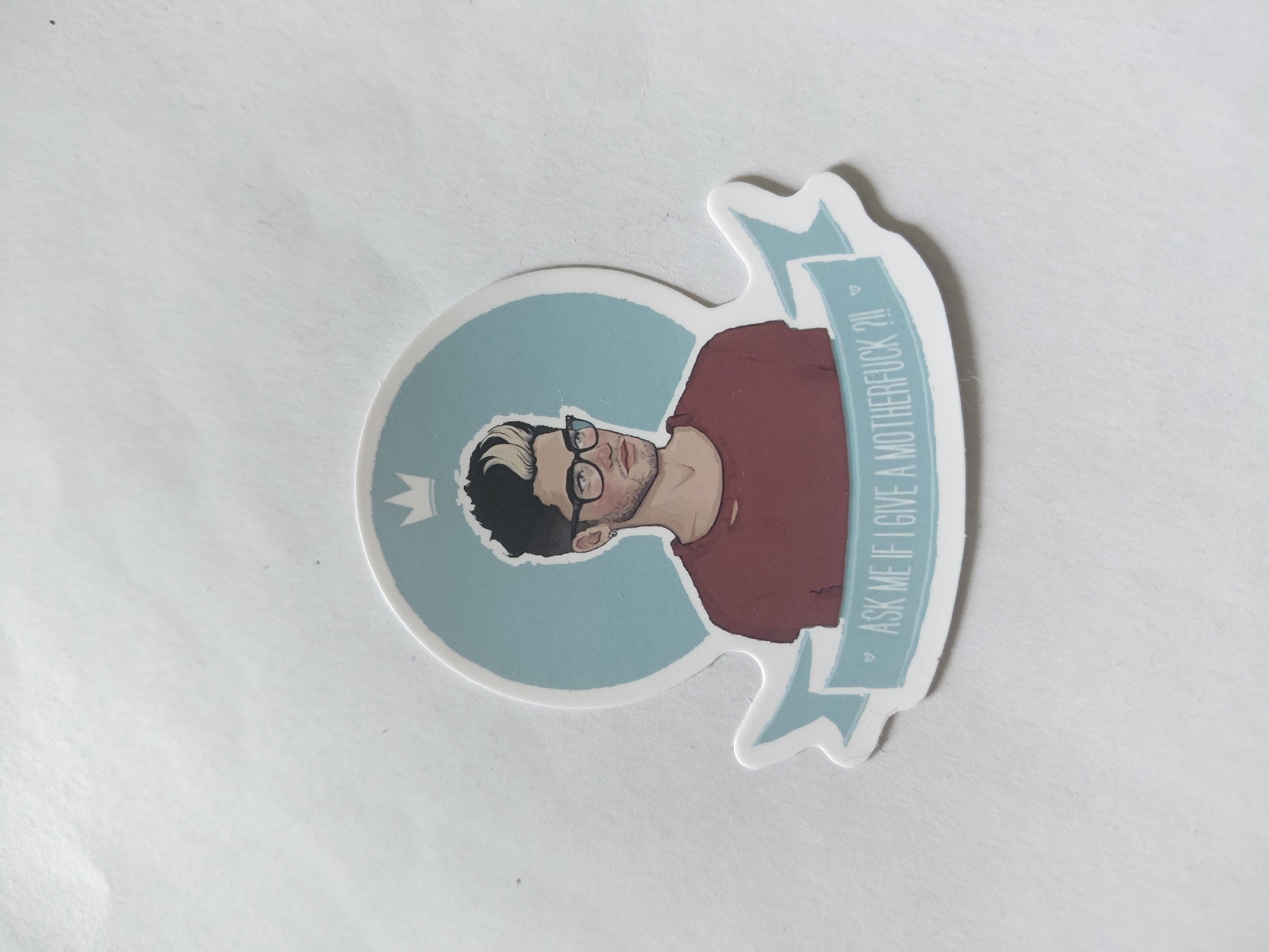 A circular sticker featuring a stylized illustration of a man wearing glasses and a maroon shirt. The man has dark hair with a blonde streak and a light beard. He is depicted from the chest up, with a light blue circle behind his head, topped with a small crown graphic. Below the man's illustration, a light blue banner is wrapped around the bottom of the circle. The banner contains white text that reads "ASK ME IF I GIVE A MOTHERFUCK ?!!" with small heart icons on either side of the question mark. The sticker is on a white, textured surface. The location is Unknown, Unknown.
