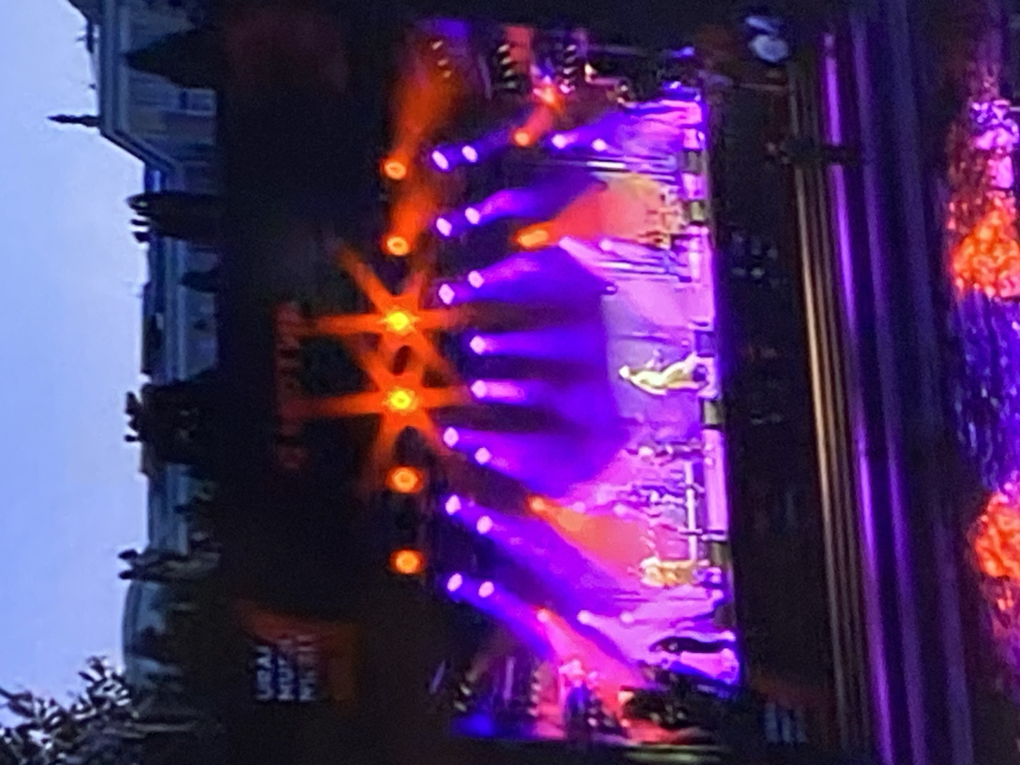 An evening outdoor concert is taking place on a stage illuminated by bright red and purple lights. A band is performing on stage, with several musicians visible. A large audience is not clearly visible, but hints of people are seen in the foreground and background. In the distance, a building with architectural details suggestive of a palace or government building is visible against a twilight sky. The text "URAL MUSIC NIGHT" is visible on a banner or sign in the upper left quadrant. The event appears to be held at a venue that includes a body of water or reflective surface in the foreground, which is also lit by the stage lights.