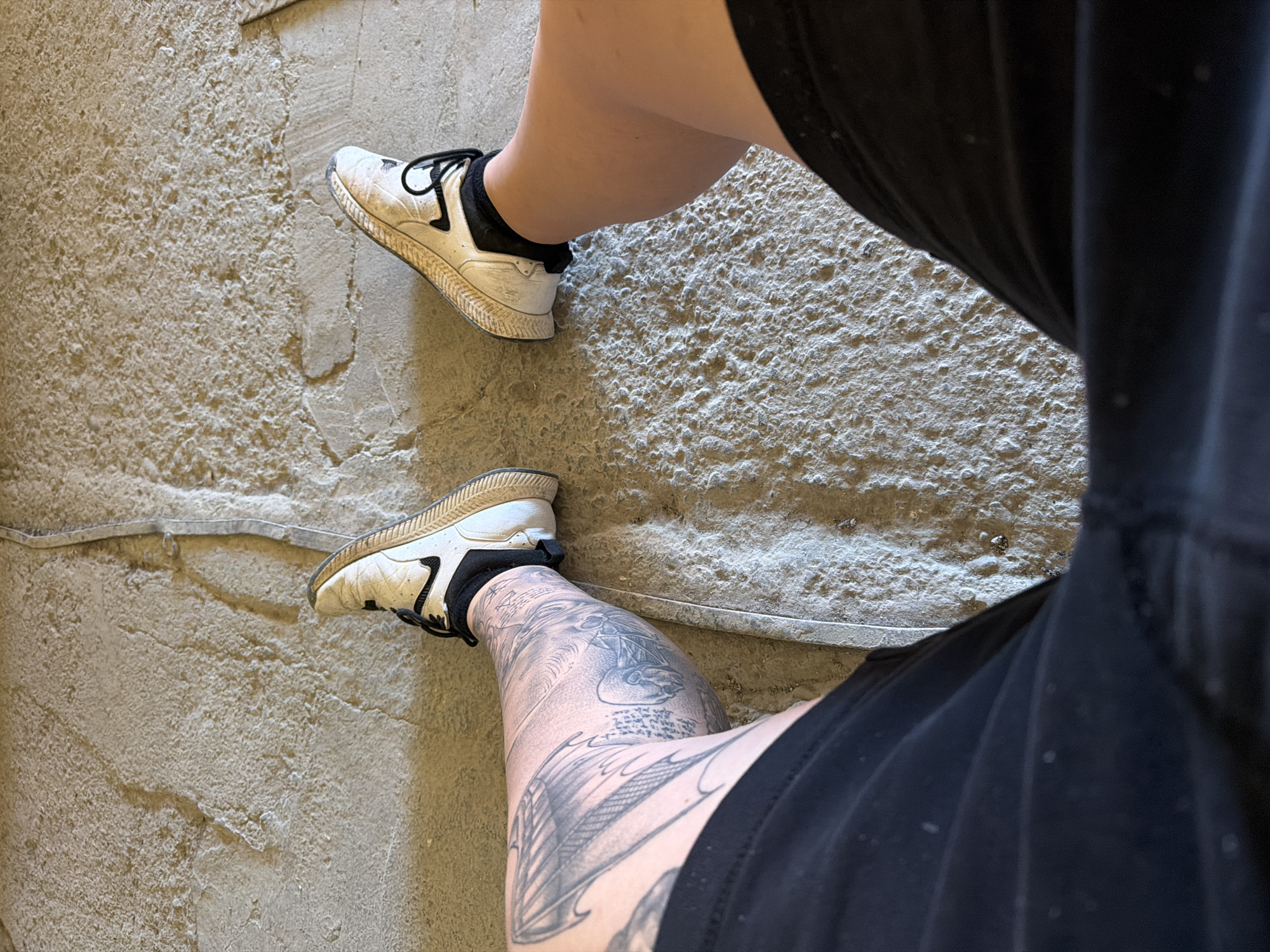 The image shows a person sitting on a rough, textured, light-colored ground, possibly concrete or a similar material. The person is wearing black shorts and white athletic shoes with black laces. The left leg, visible from the knee down, has extensive tattoos covering the thigh and calf. The right leg is also visible from the knee down. The ground appears to be outdoors, possibly in an undeveloped or construction area. There is a thin, lighter-colored strip running diagonally across the ground, which may be a seam or a piece of tape. The scene does not provide any specific visual cues to indicate a precise location within Dolno Ablanovo, Bulgaria.